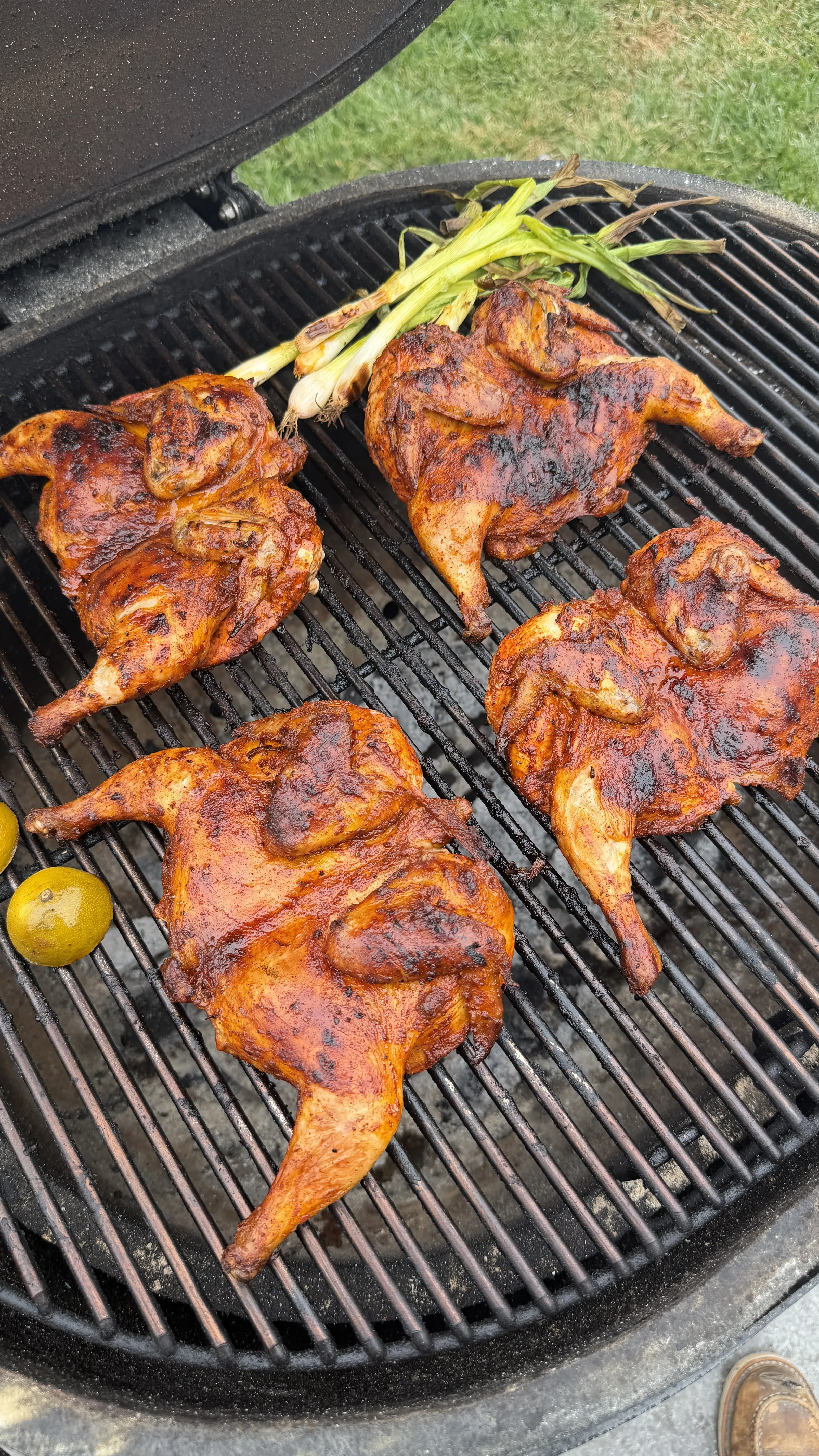 Four spatchcocked pollo asado Cornish hens grilling on a charcoal grill with green onions and limes