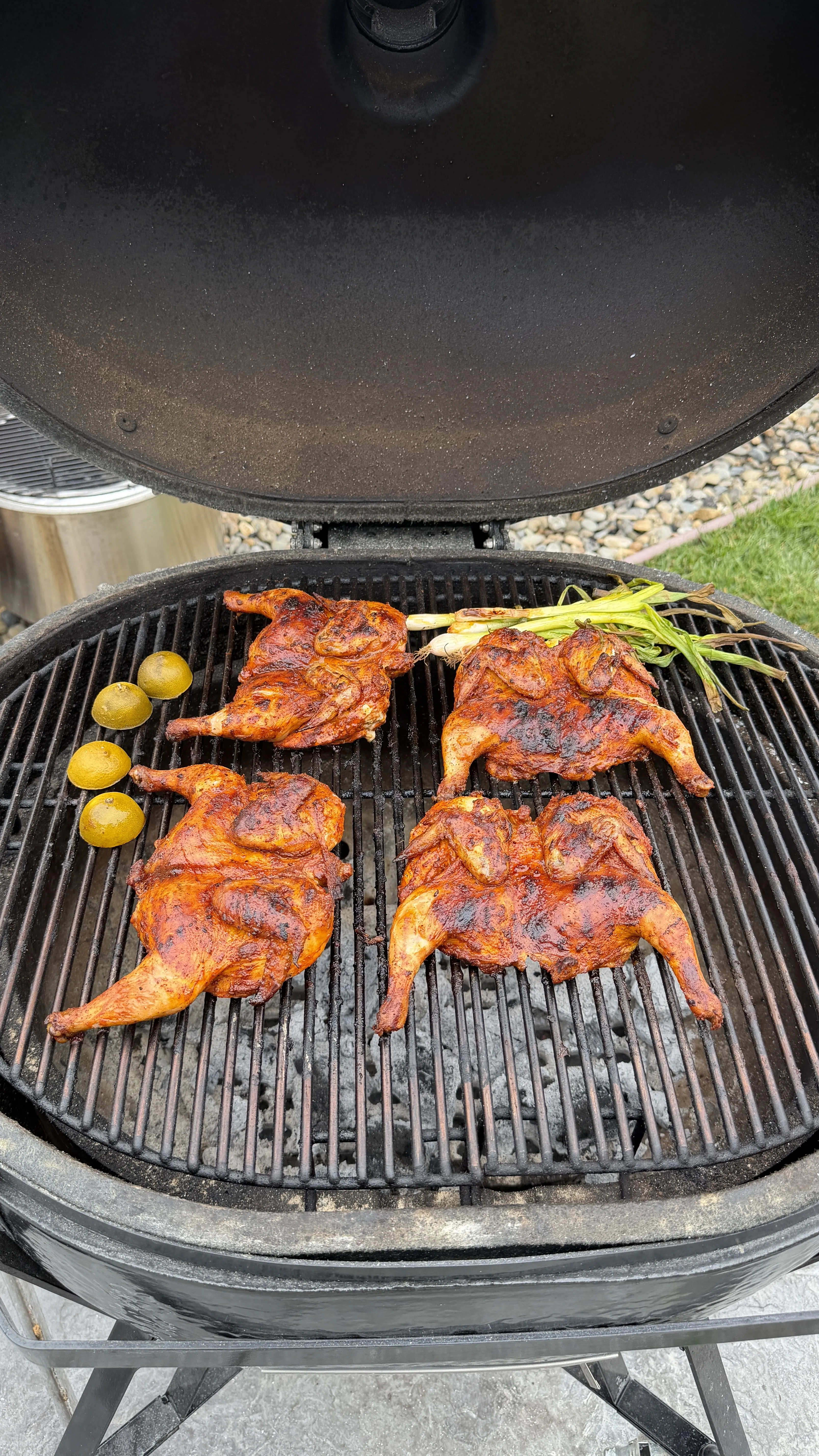 Four spatchcocked pollo asado Cornish hens grilling on a charcoal kamado grill with limes and green onions