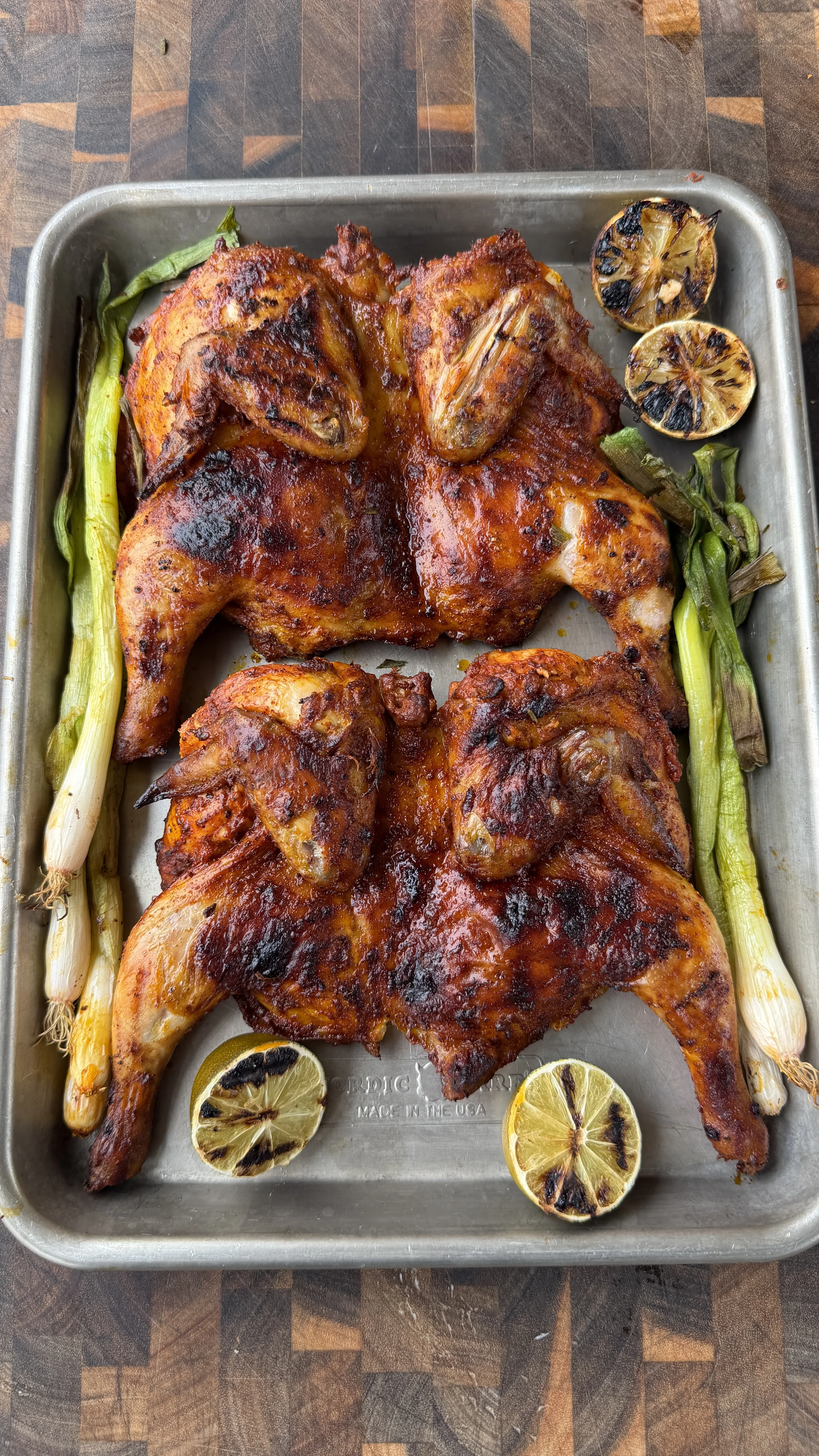 Two spatchcocked pollo asado Cornish hens on a sheet pan with charred limes and grilled green onions