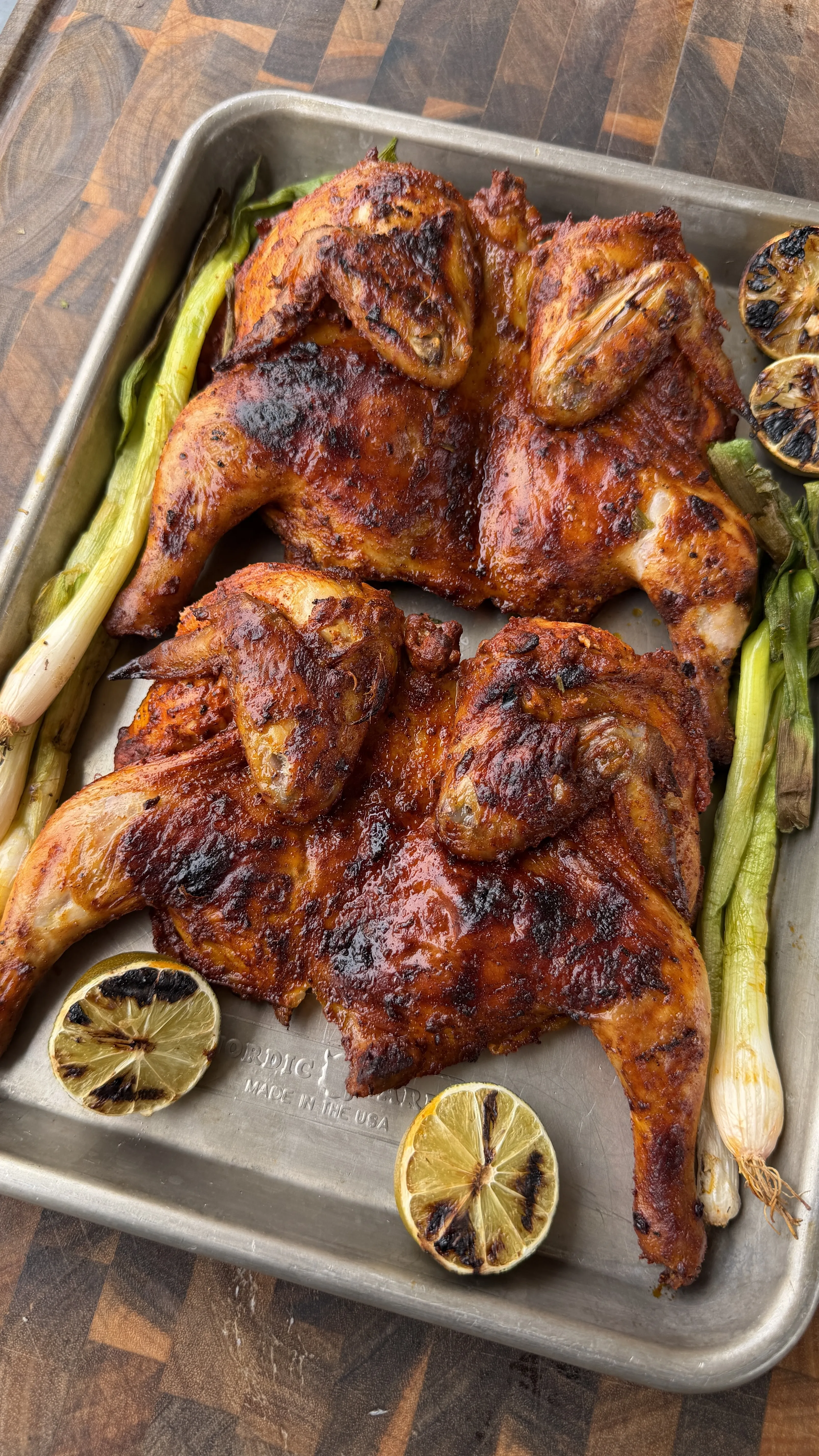 Two spatchcocked pollo asado style Cornish hens on a sheet pan with charred limes and grilled green onions