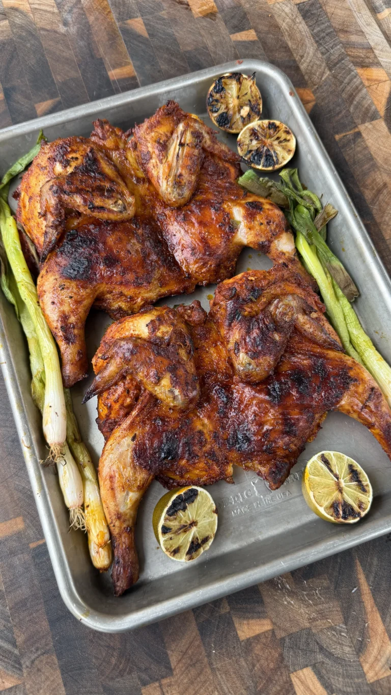 Two spatchcocked pollo asado cornish hens on a sheet pan with charred lime halves and grilled green onions