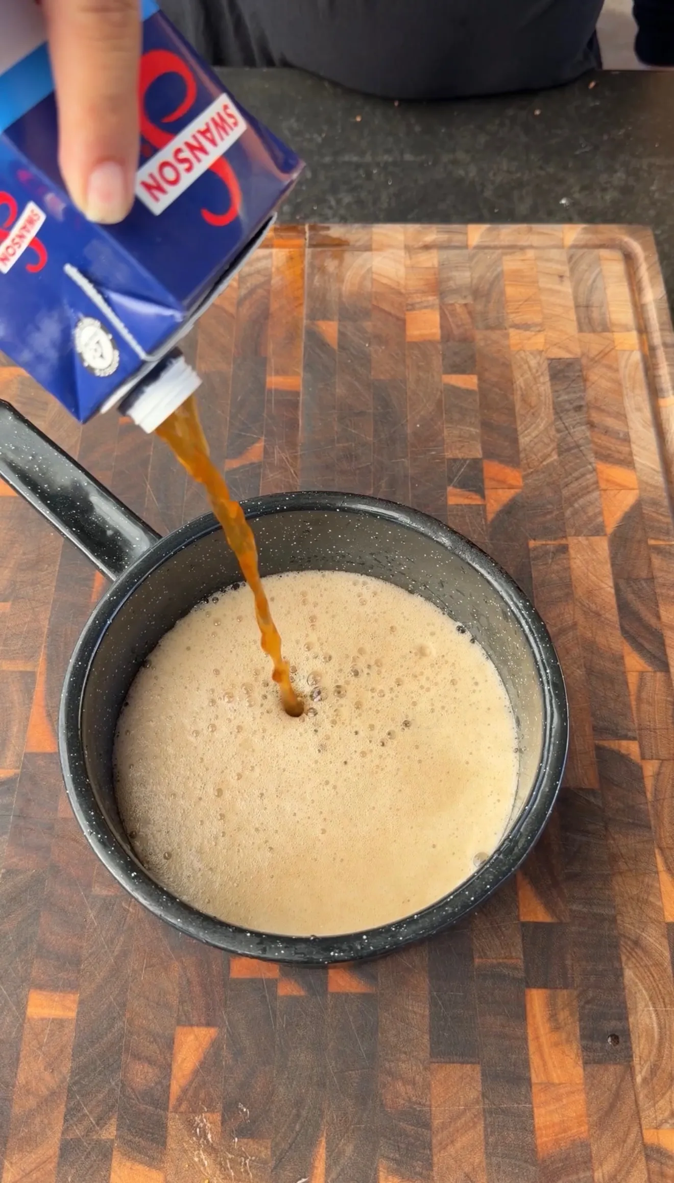 Pouring Swanson chicken broth from carton into a dark saucepan with creamy liquid for smoked turkey legs