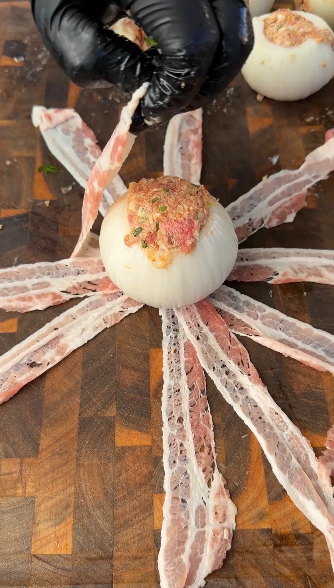 Raw onion stuffed with seasoned ground meat, surrounded by strips of bacon on a wooden cutting board