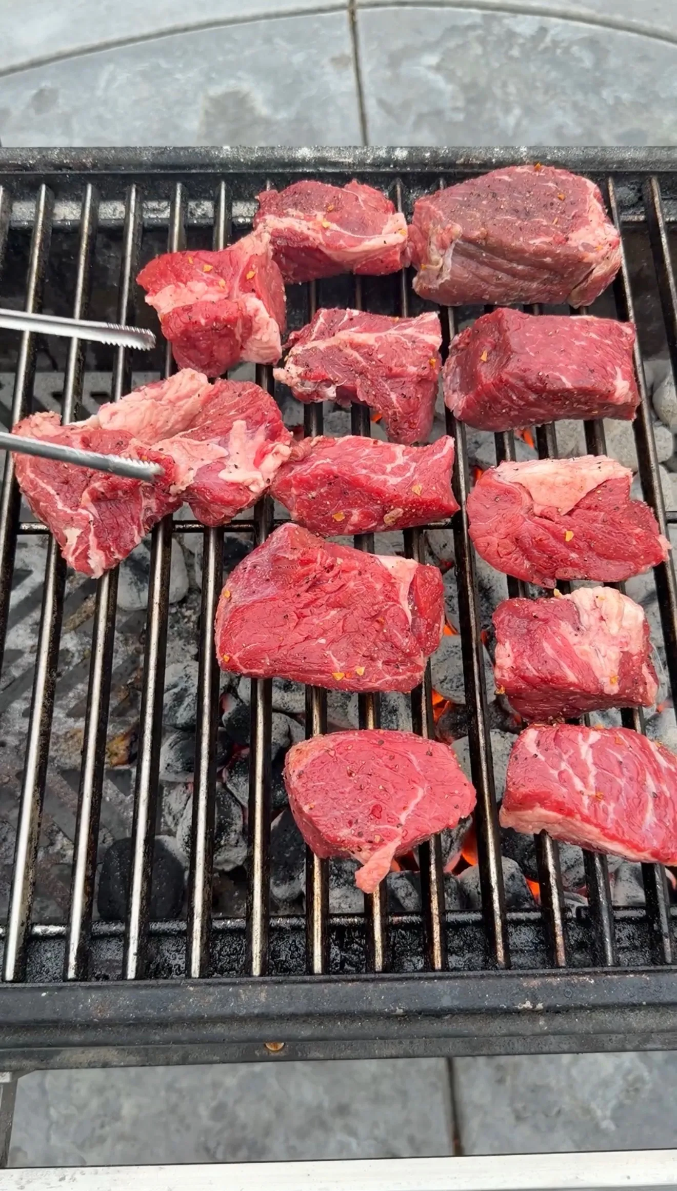 Seasoned raw beef chunks for birria tacos cooking over hot charcoal grill grates with tongs