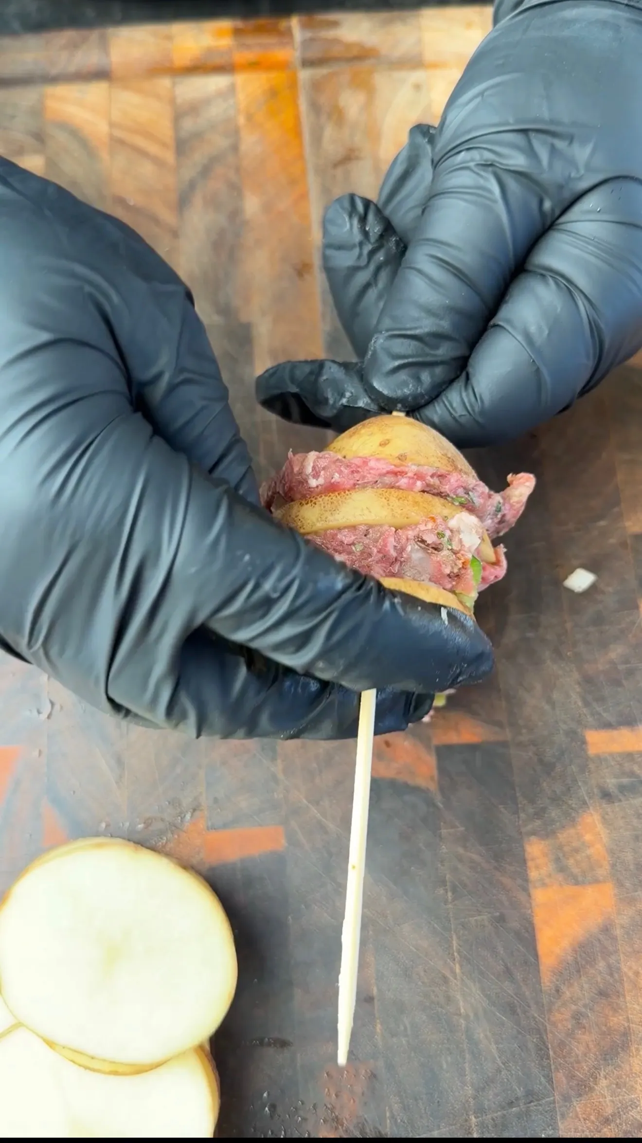 Gloved hands pressing seasoned raw ground beef into sliced potato on a wooden skewer for Cajun kebabs