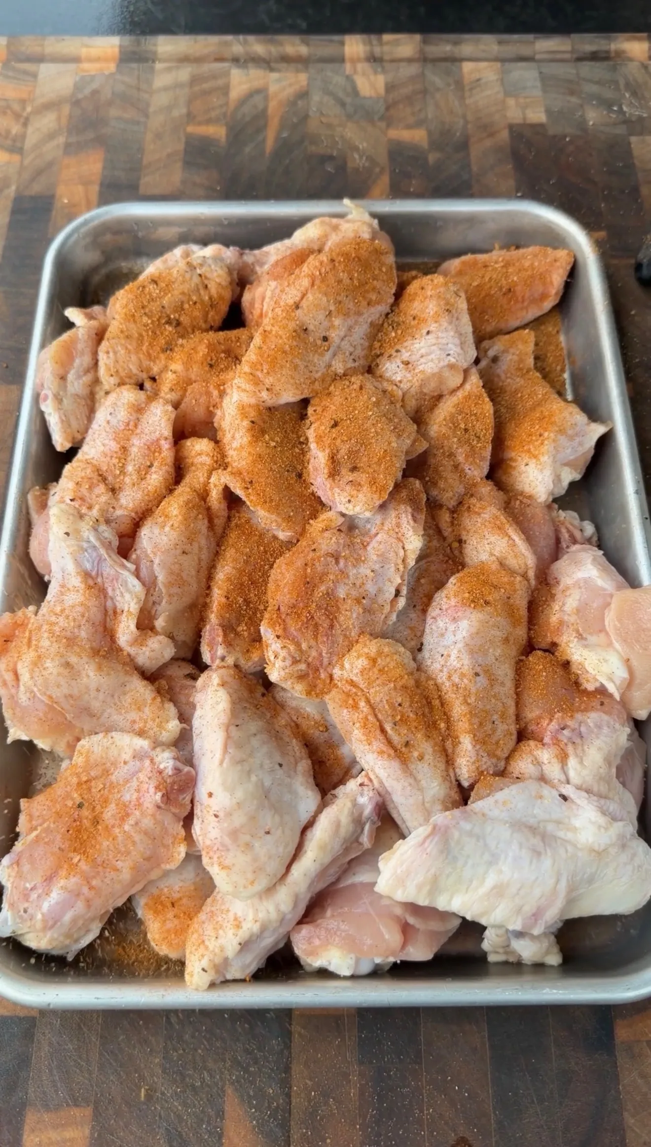Raw chicken wings coated in orange-red spice rub in a stainless steel baking pan on a wooden cutting board