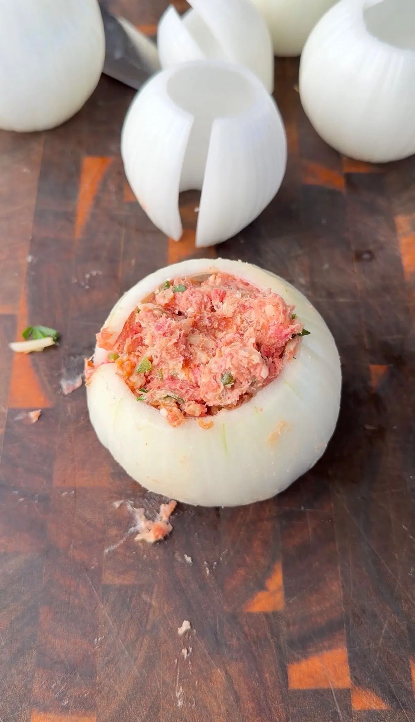 Hollowed white onion filled with seasoned ground meat mixture on a wooden cutting board, ready to be wrapped in bacon