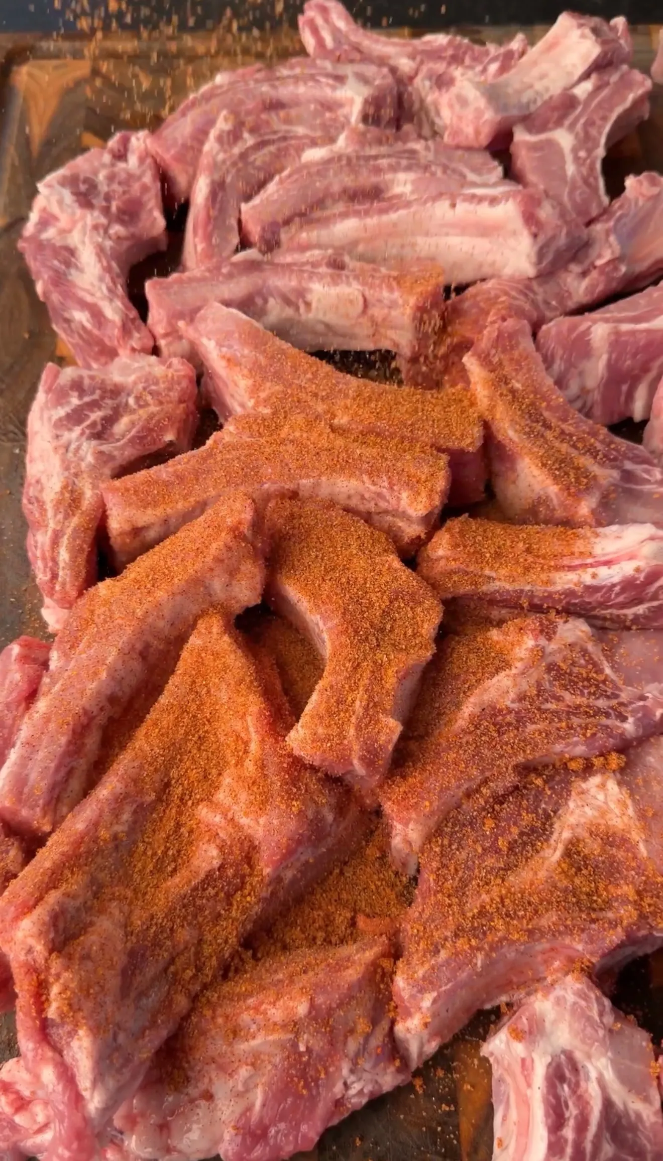 Raw pork ribs being seasoned with a red-orange spice dry rub on a wooden cutting board