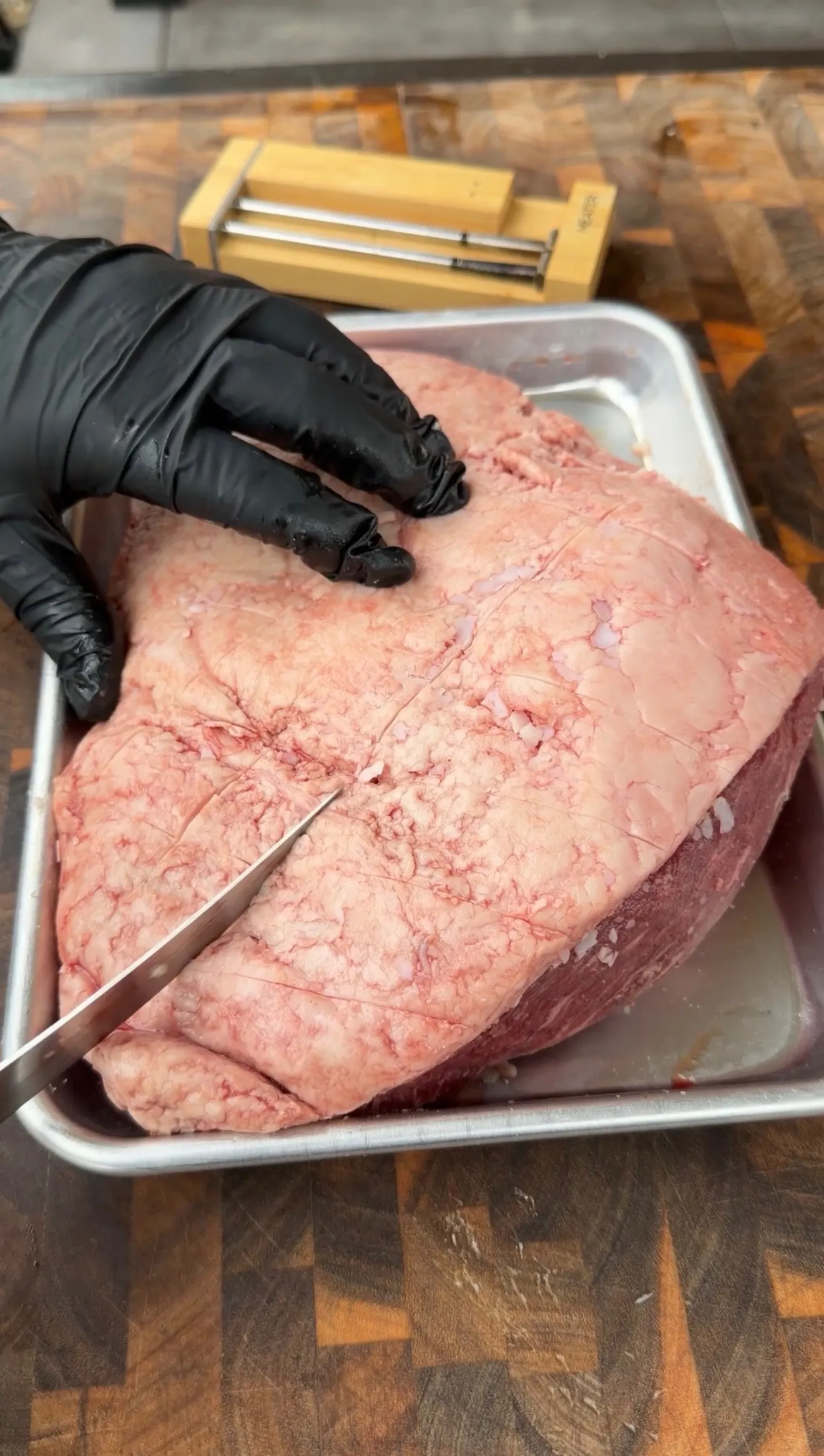 Gloved hands trimming fat cap on a large raw picanha roast in a metal pan on a butcher block