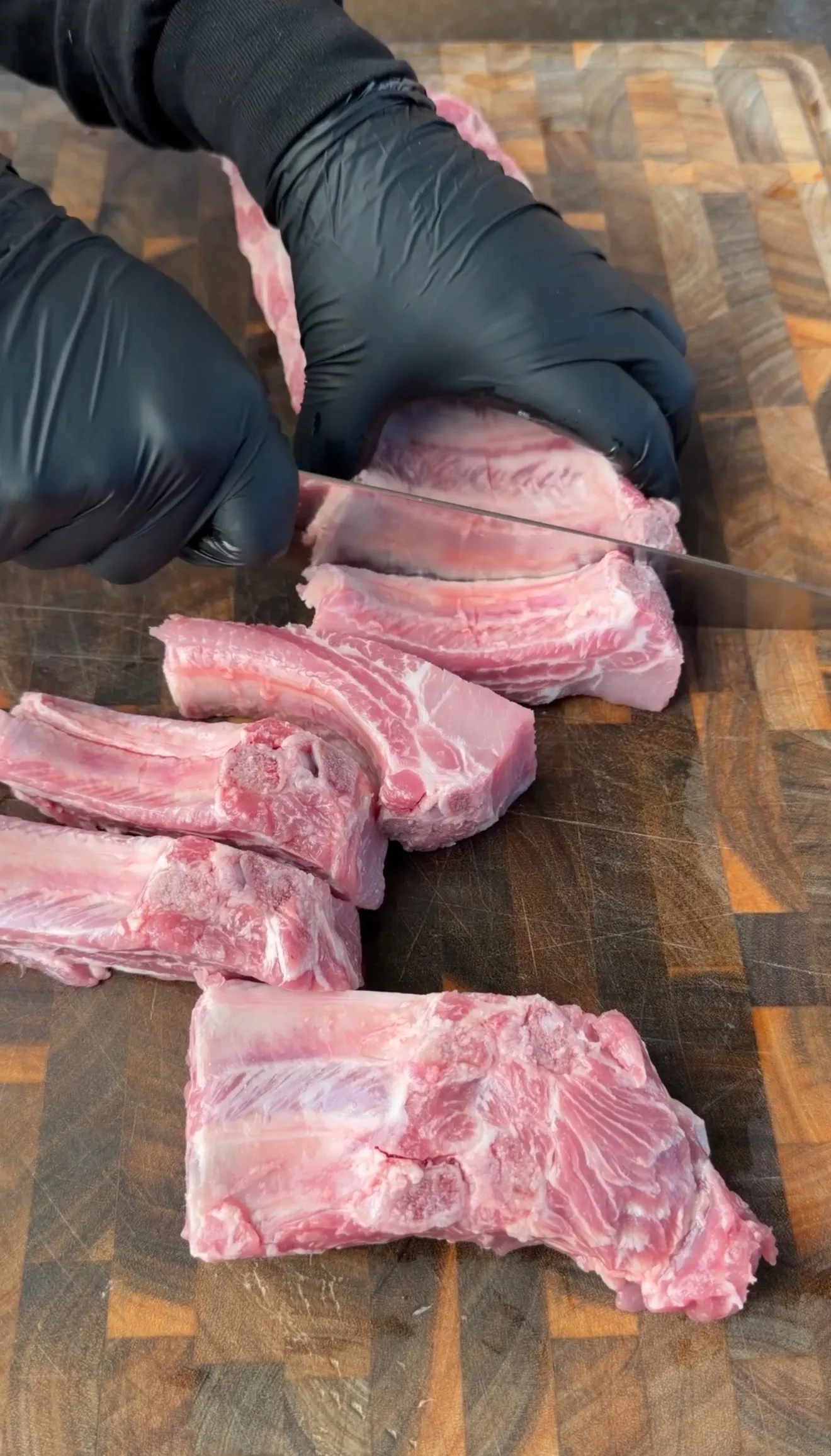 Gloved hands slicing raw pork ribs into individual portions on a wooden end-grain cutting board