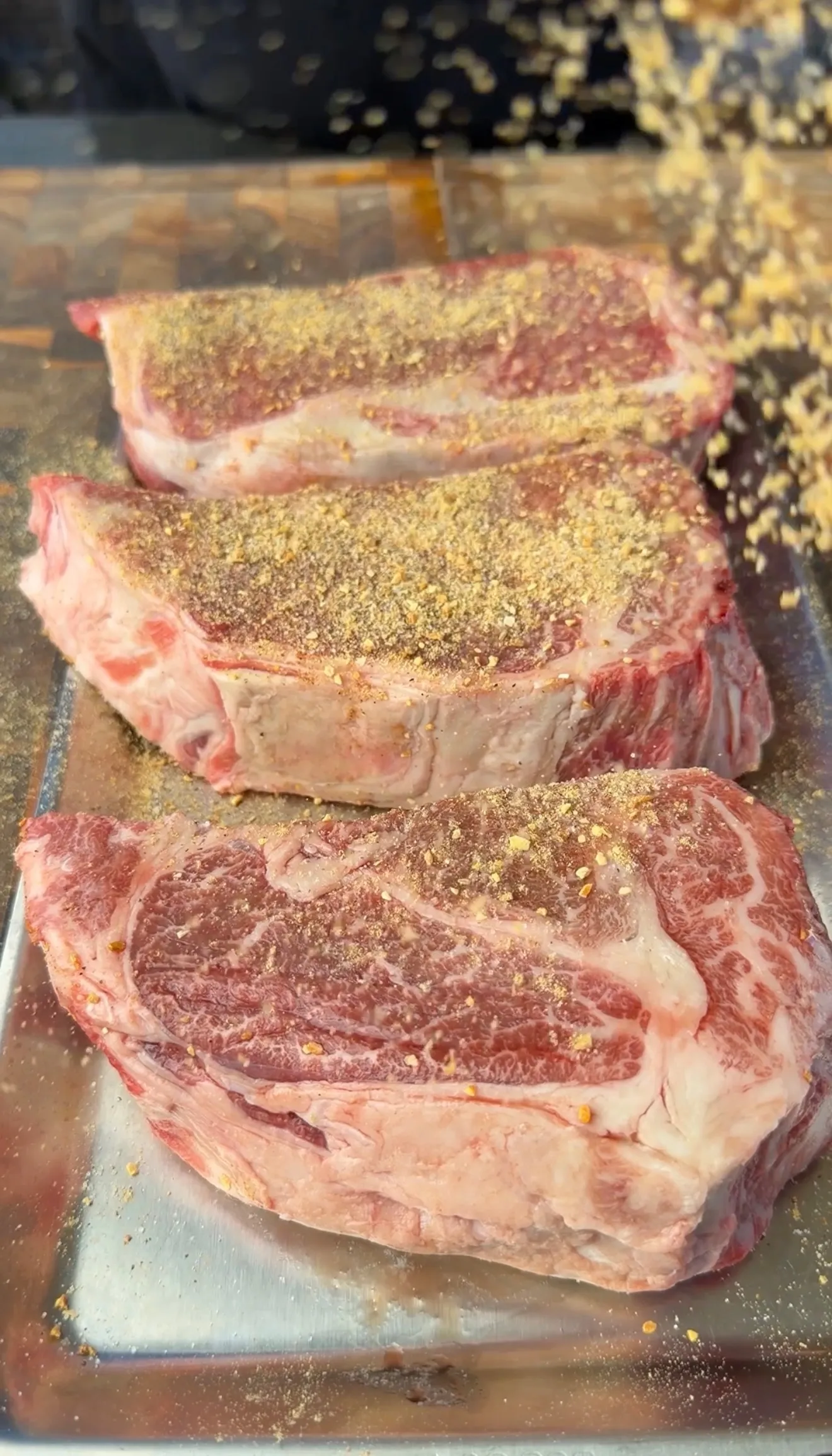 Three thick raw ribeye steaks generously coated with seasoning rub on a metal baking sheet