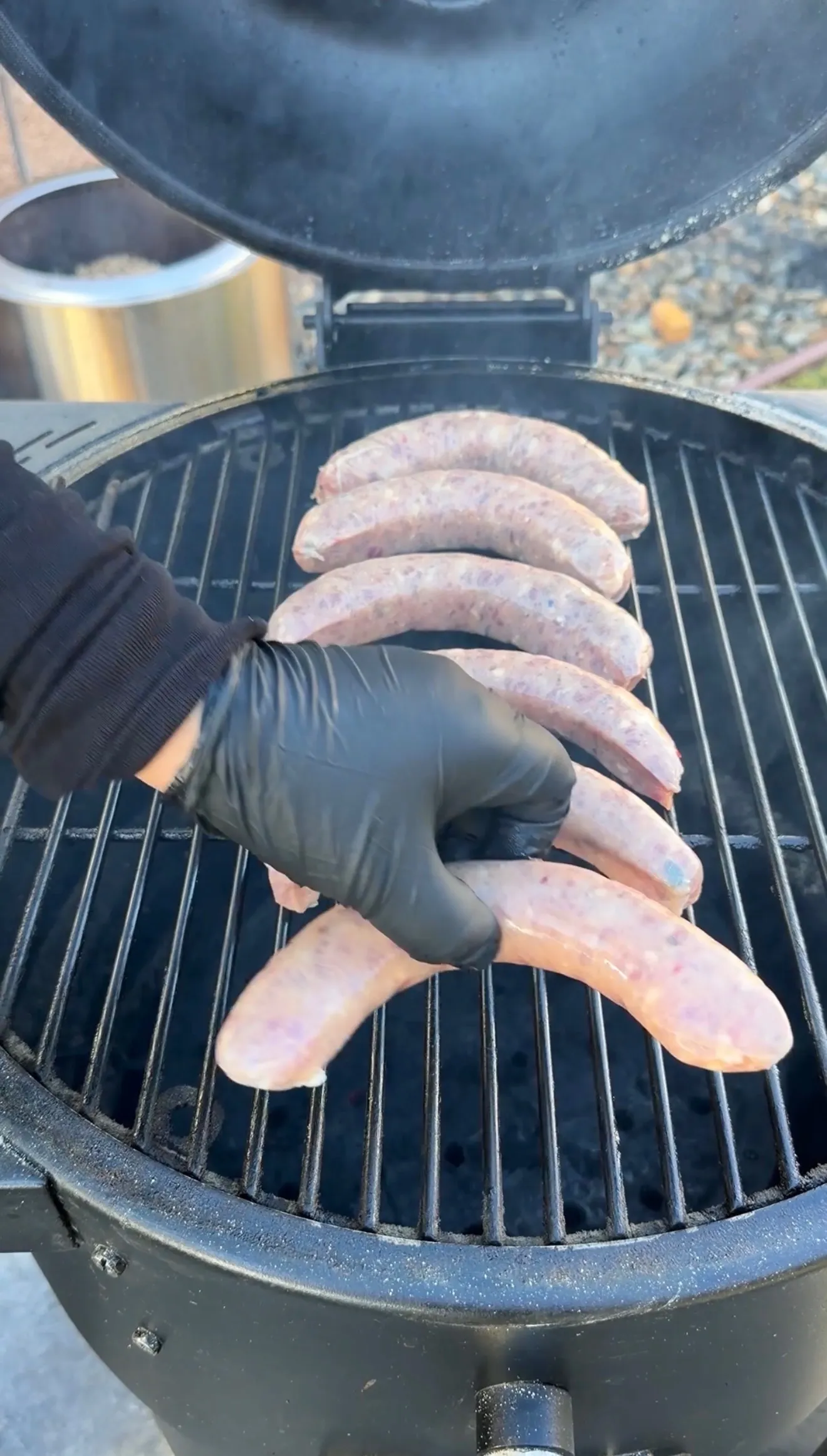 Gloved hand placing raw sausage links onto a round charcoal grill for smoked sausage breakfast skillet
