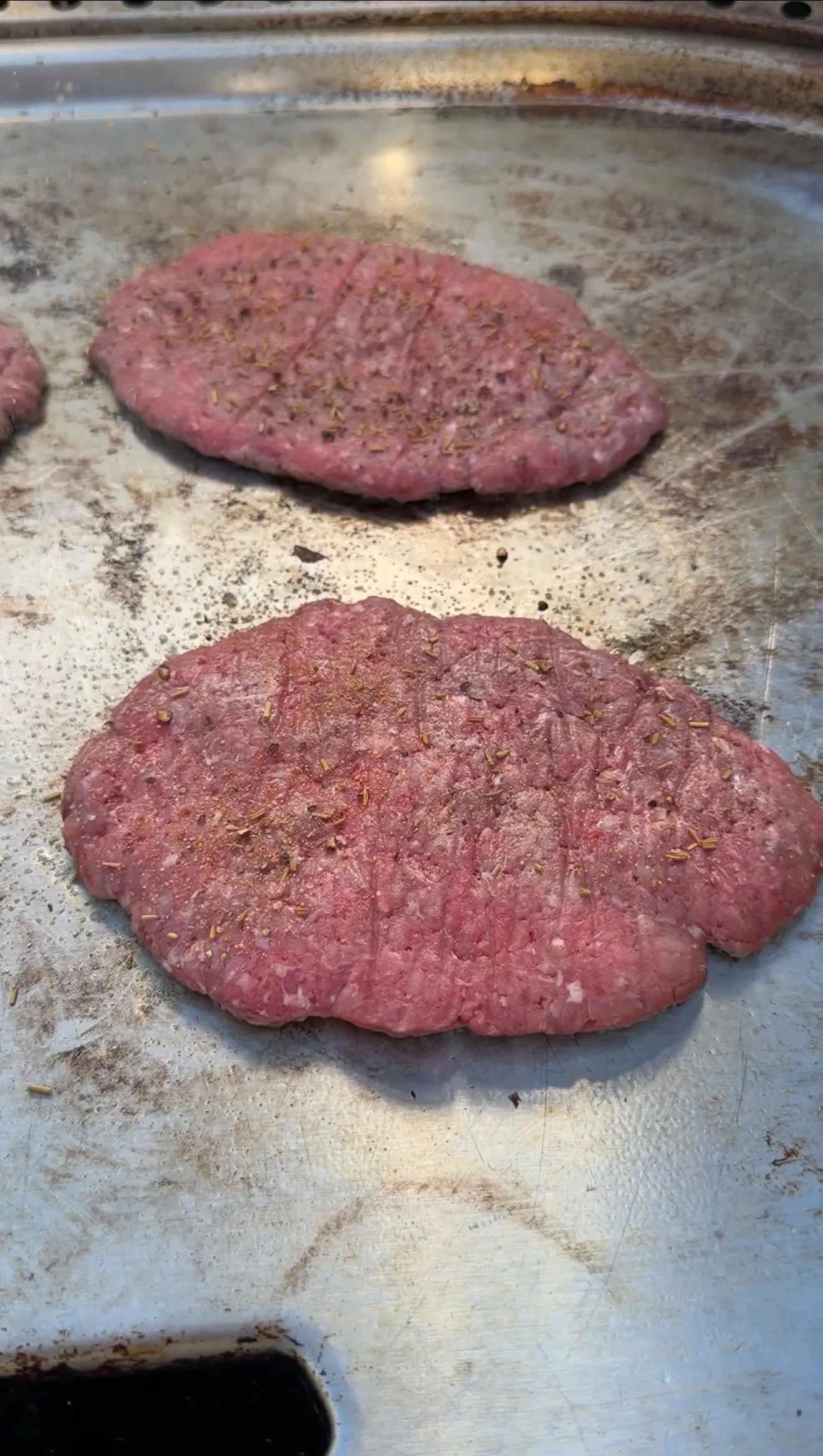 Two raw ground beef burger patties seasoned with herbs and spices on a metal baking sheet