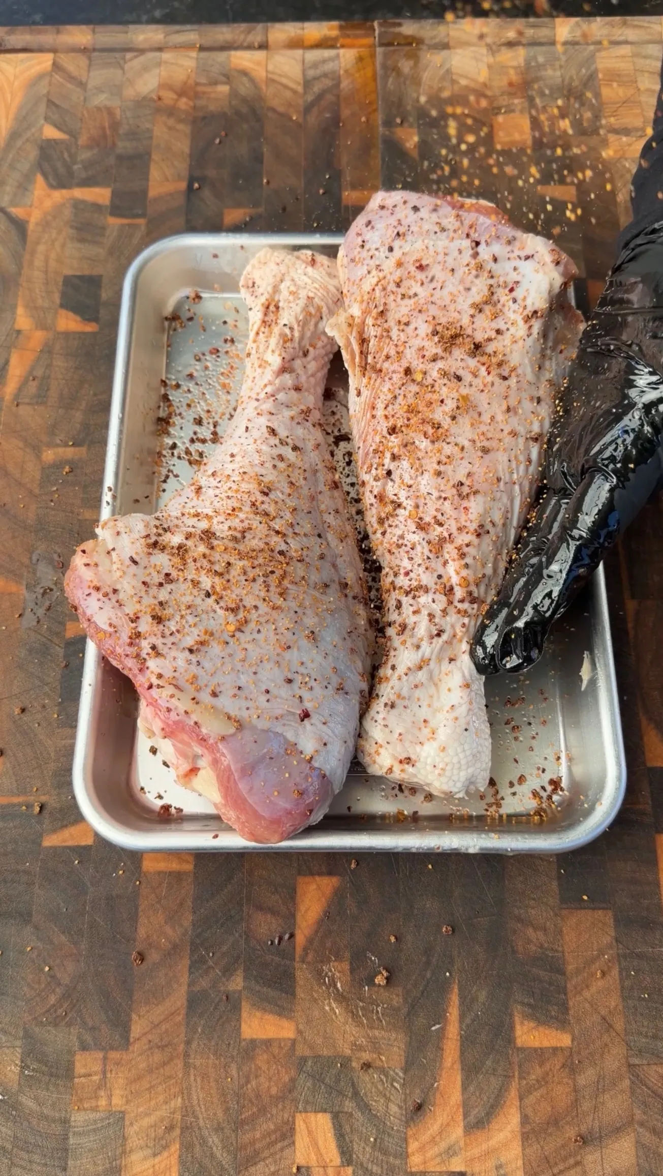 Two raw turkey legs coated in dry rub seasoning on a metal sheet pan, ready for smoking