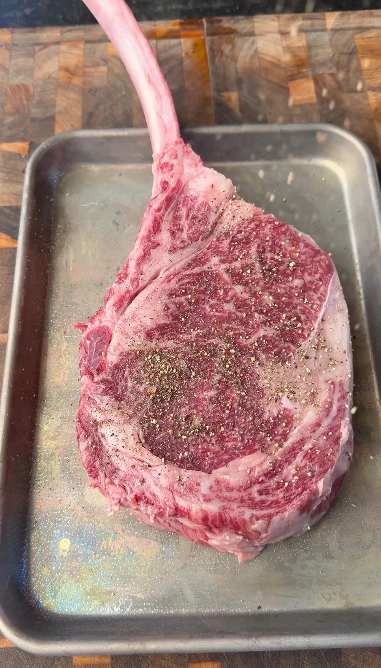 Raw tomahawk ribeye steak seasoned with salt and pepper on a stainless steel tray, ready to cook