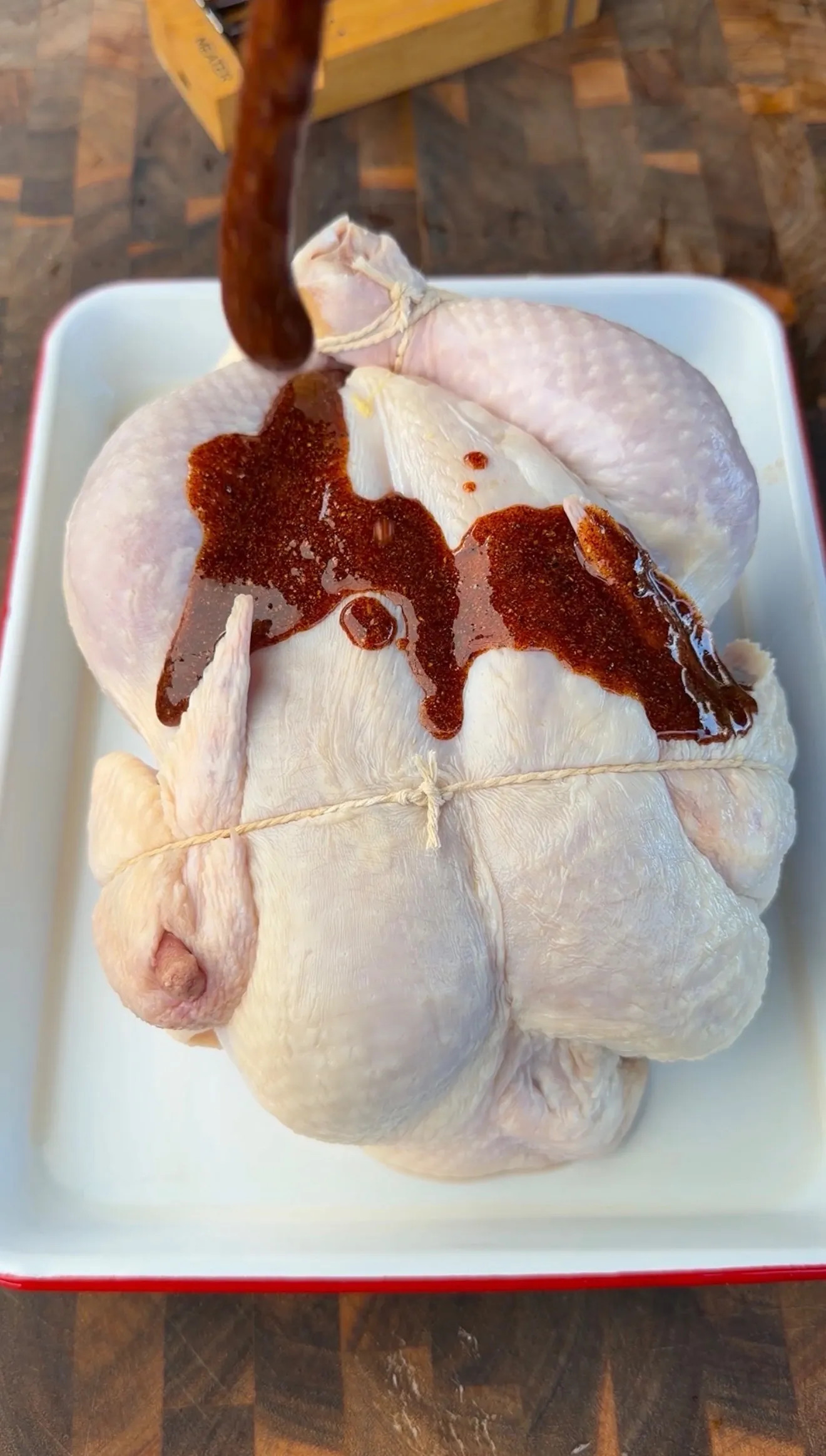 Raw trussed whole chicken in white baking dish being coated with honey lemon pepper marinade sauce