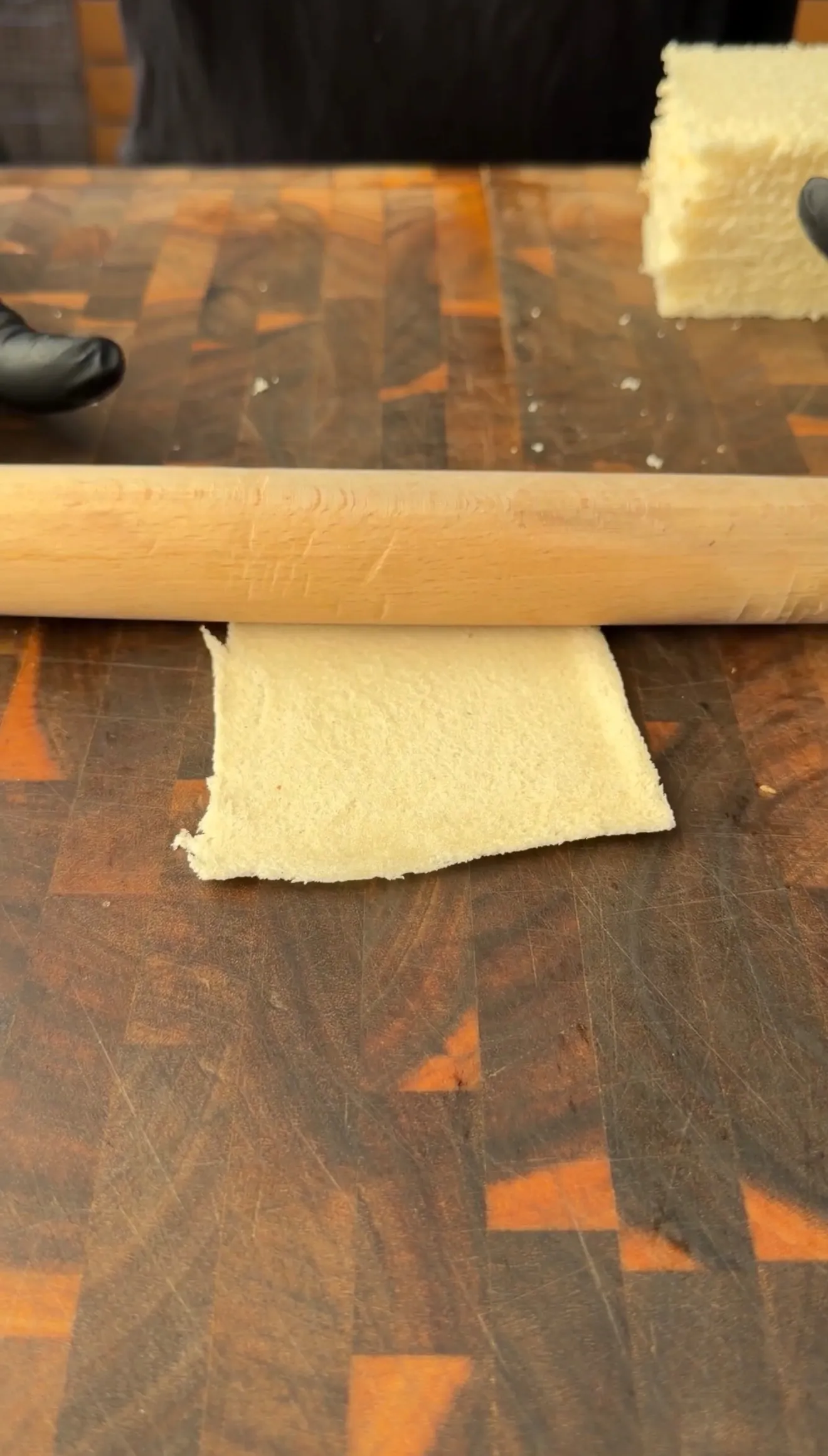 A rolling pin flattening a slice of white bread on a wooden cutting board for cheesesteak stuffed pockets