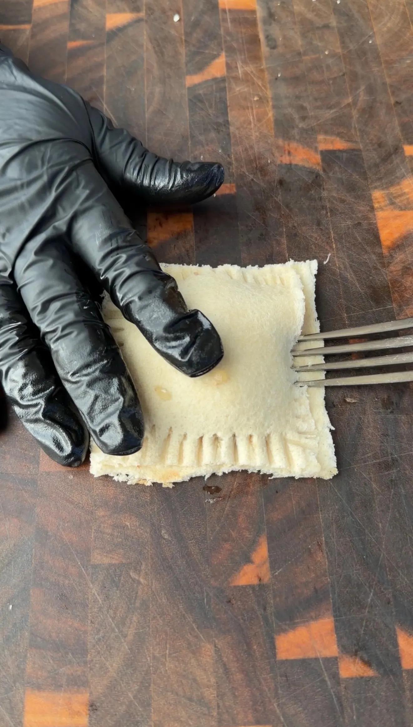 Black-gloved hand pressing fork tines to seal edges of a cheesesteak stuffed bread pocket on wooden cutting board