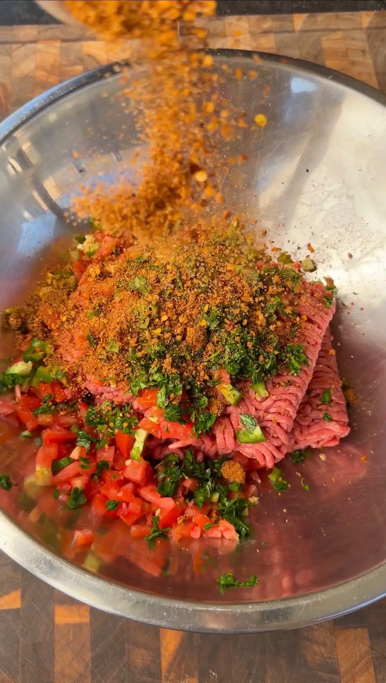 Ground beef with diced tomatoes, fresh herbs, and spices being added to a stainless steel mixing bowl