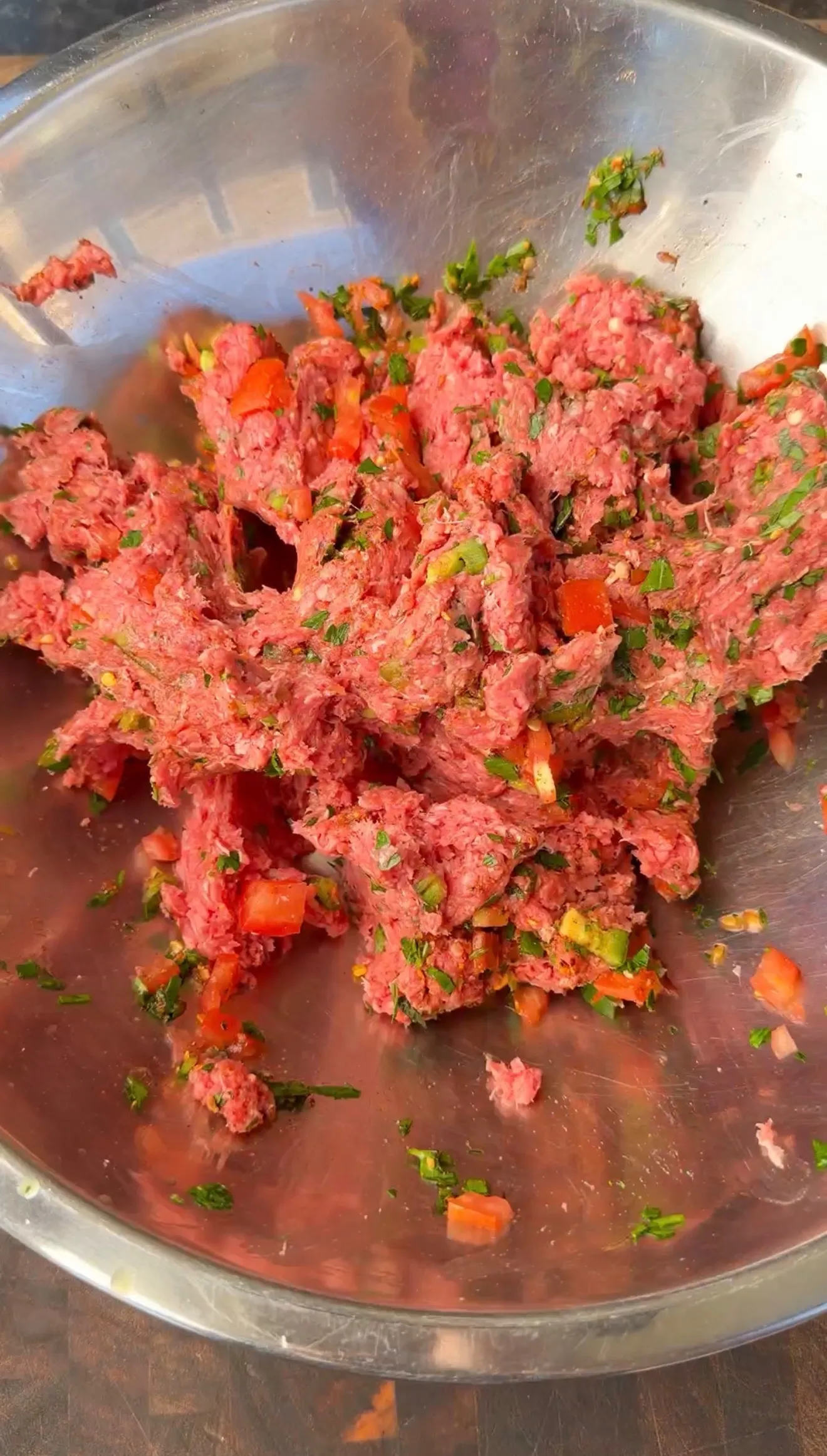 Raw seasoned ground beef mixed with diced red peppers and fresh herbs in a stainless steel bowl