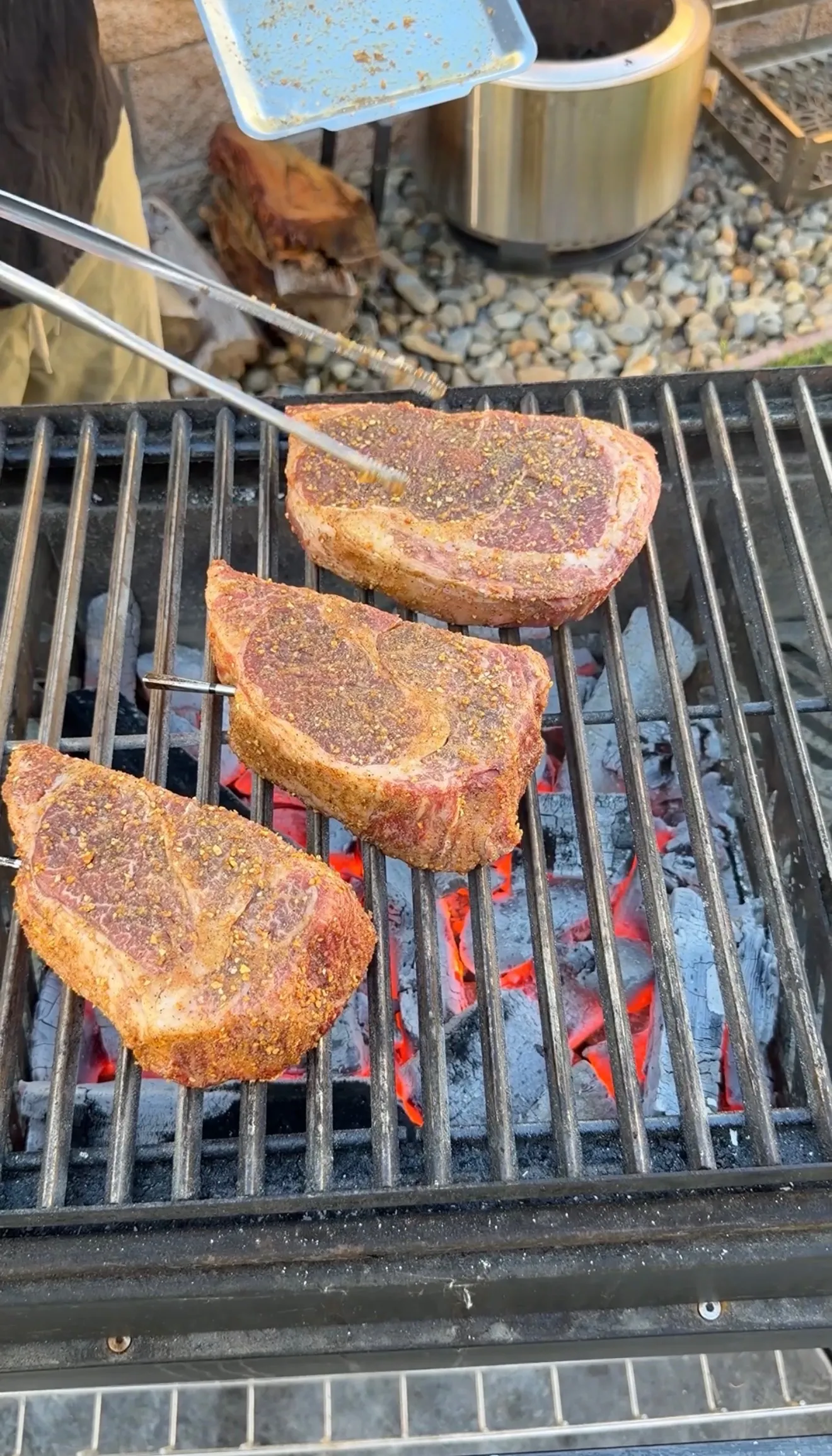 Three thick seasoned ribeye steaks grilling over hot charcoal with tongs placing one on the grate