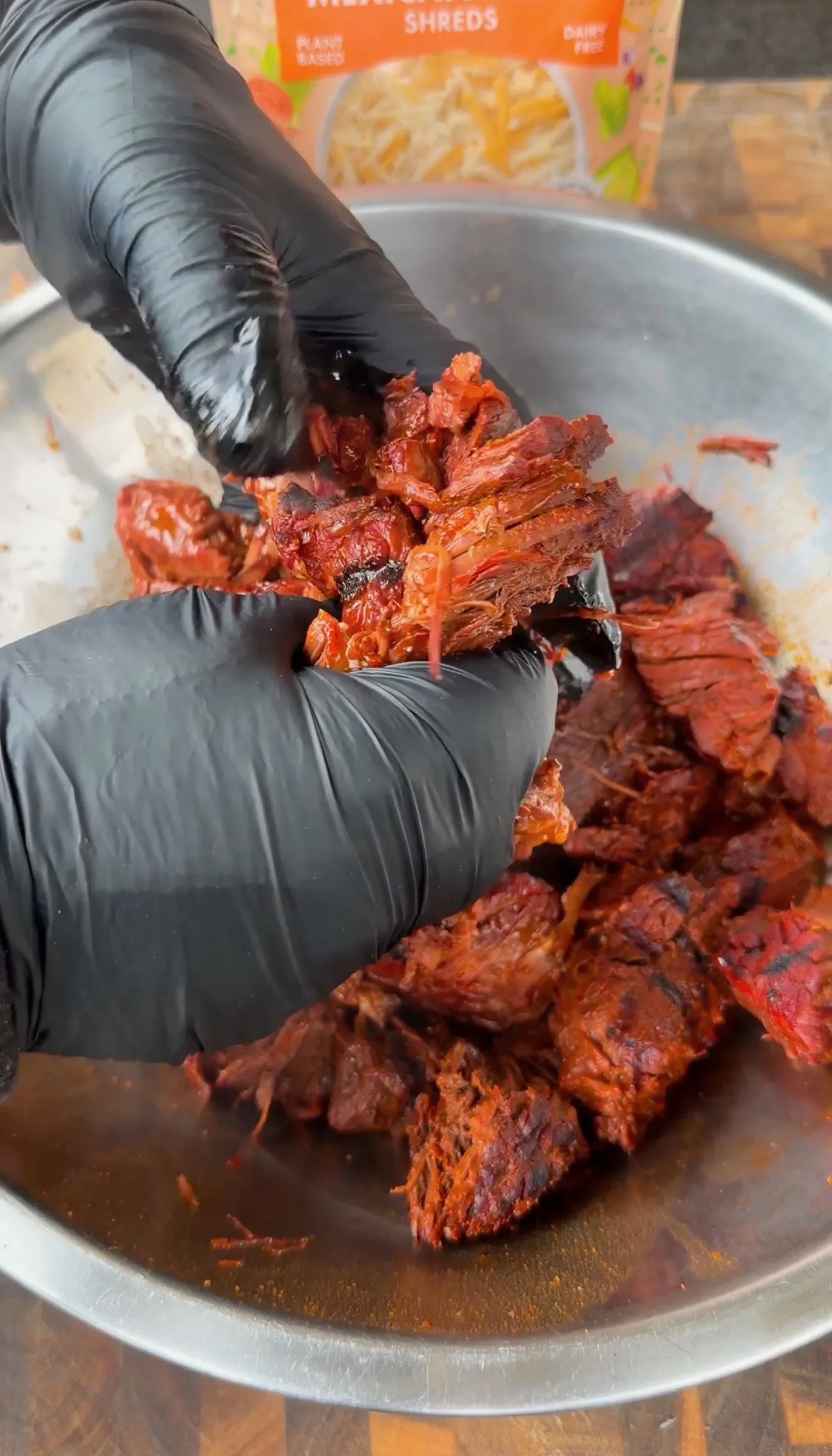 Gloved hands pulling apart seasoned shredded birria beef in a stainless steel mixing bowl