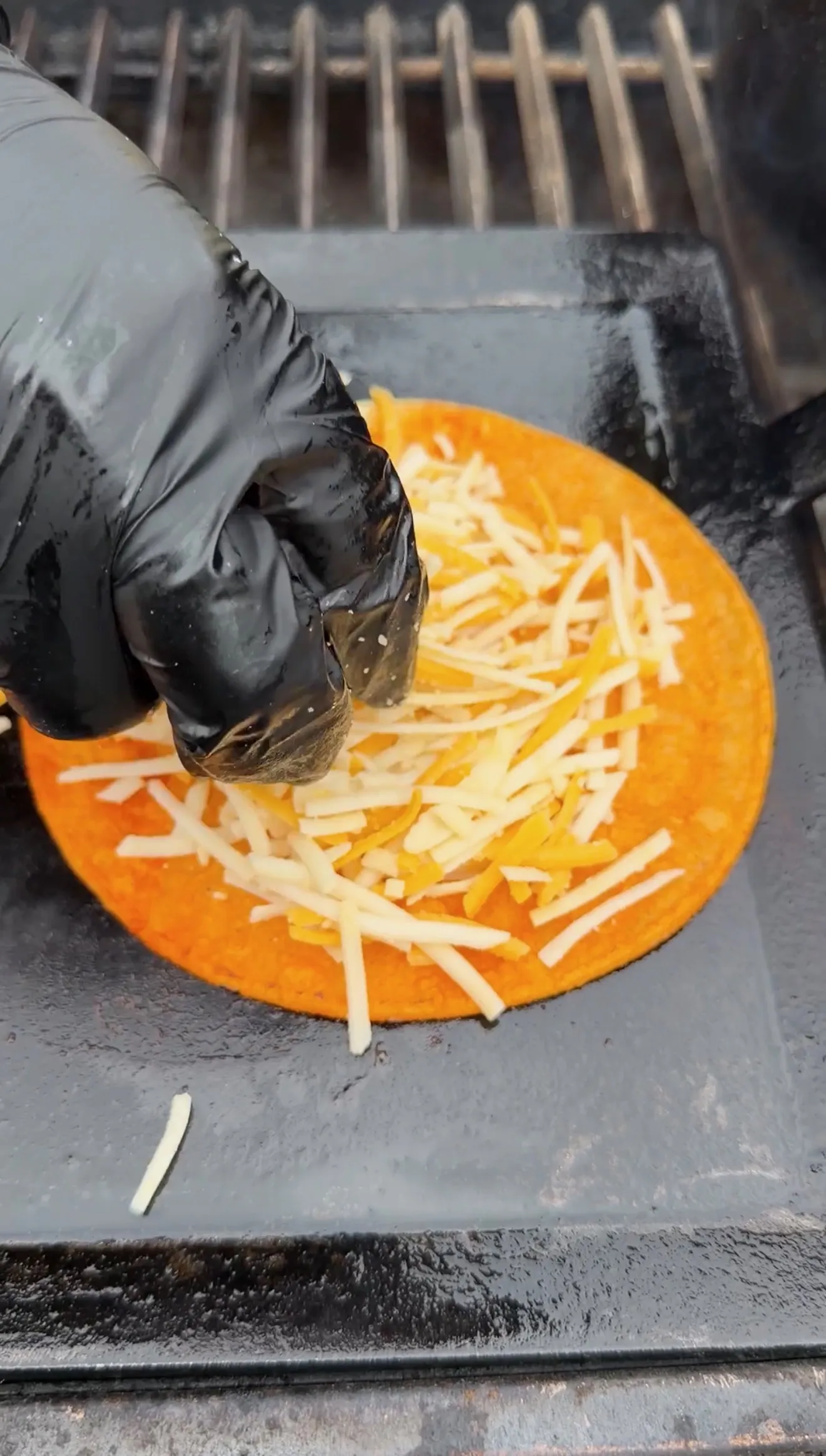 Black-gloved hand sprinkling shredded white and orange cheese onto a corn tortilla on a hot griddle