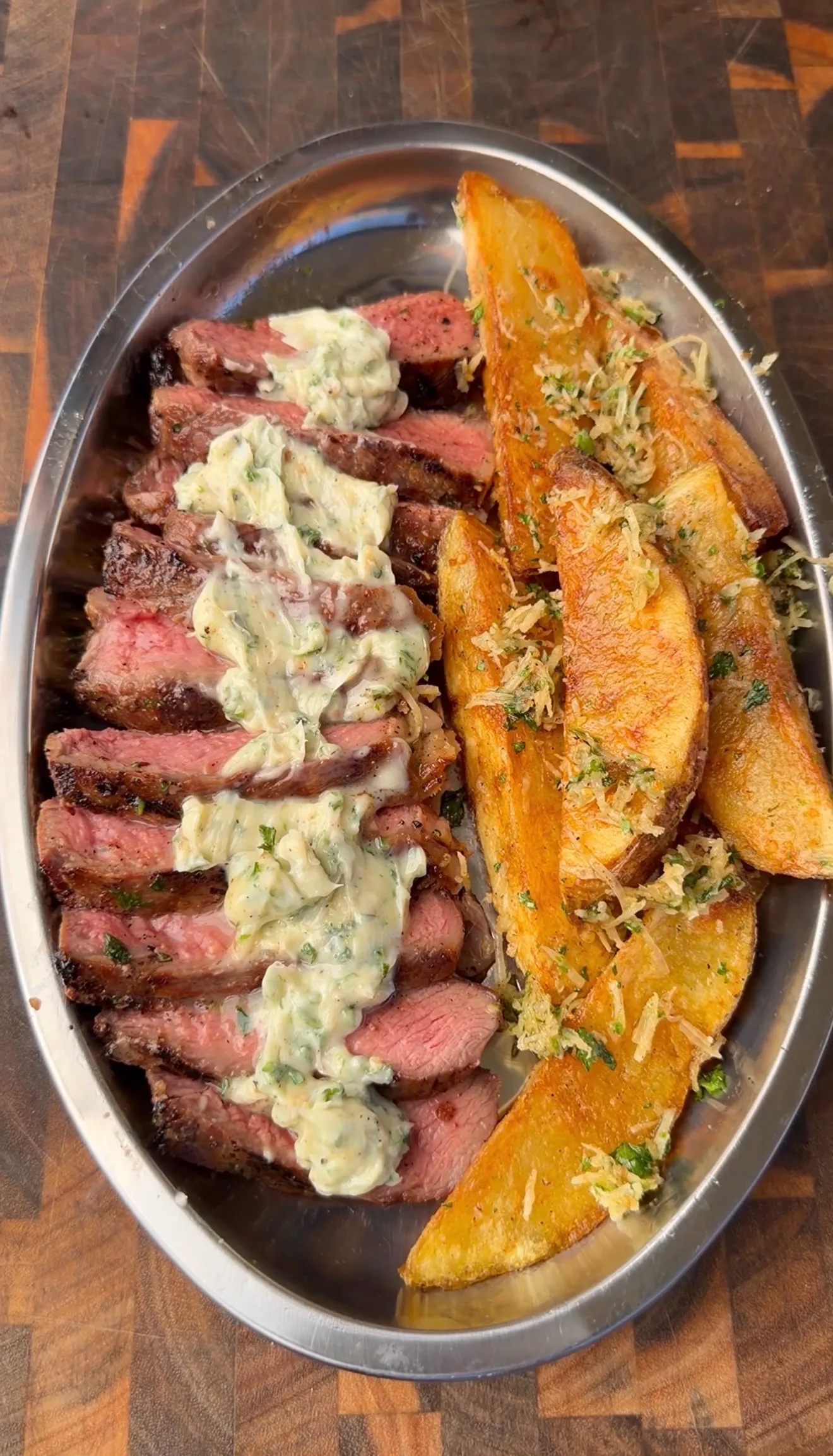 Sliced grilled ribeye steak topped with umami butter served alongside crispy Parmesan potato wedges on a silver platter