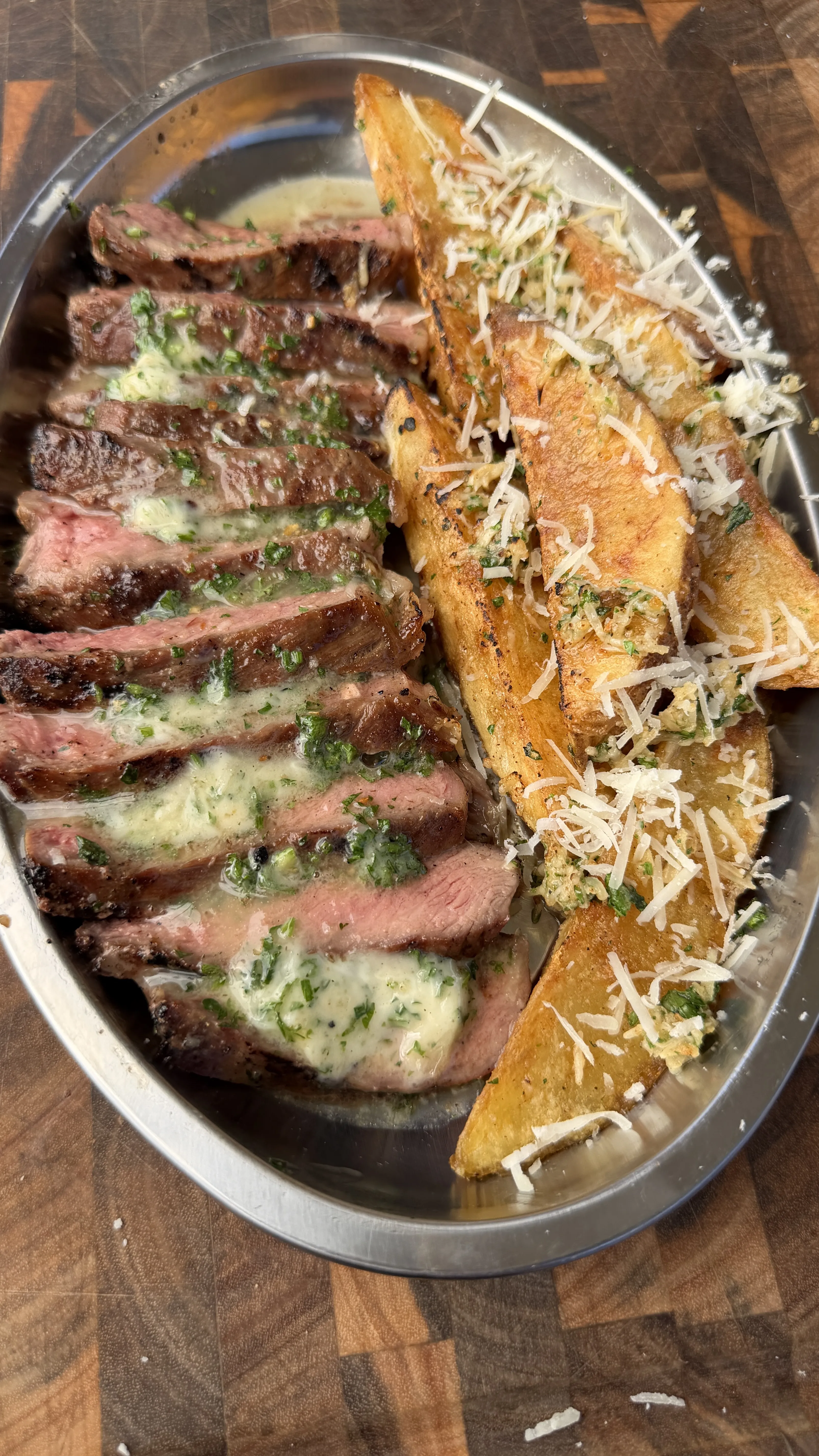 Sliced grilled ribeye steak with melted umami herb butter and crispy Parmesan potato wedges on a silver platter