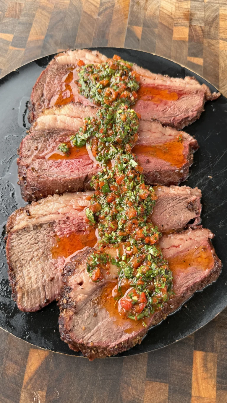 Sliced smoked picanha roast topped with red cilantro chimichurri sauce on a black oval plate
