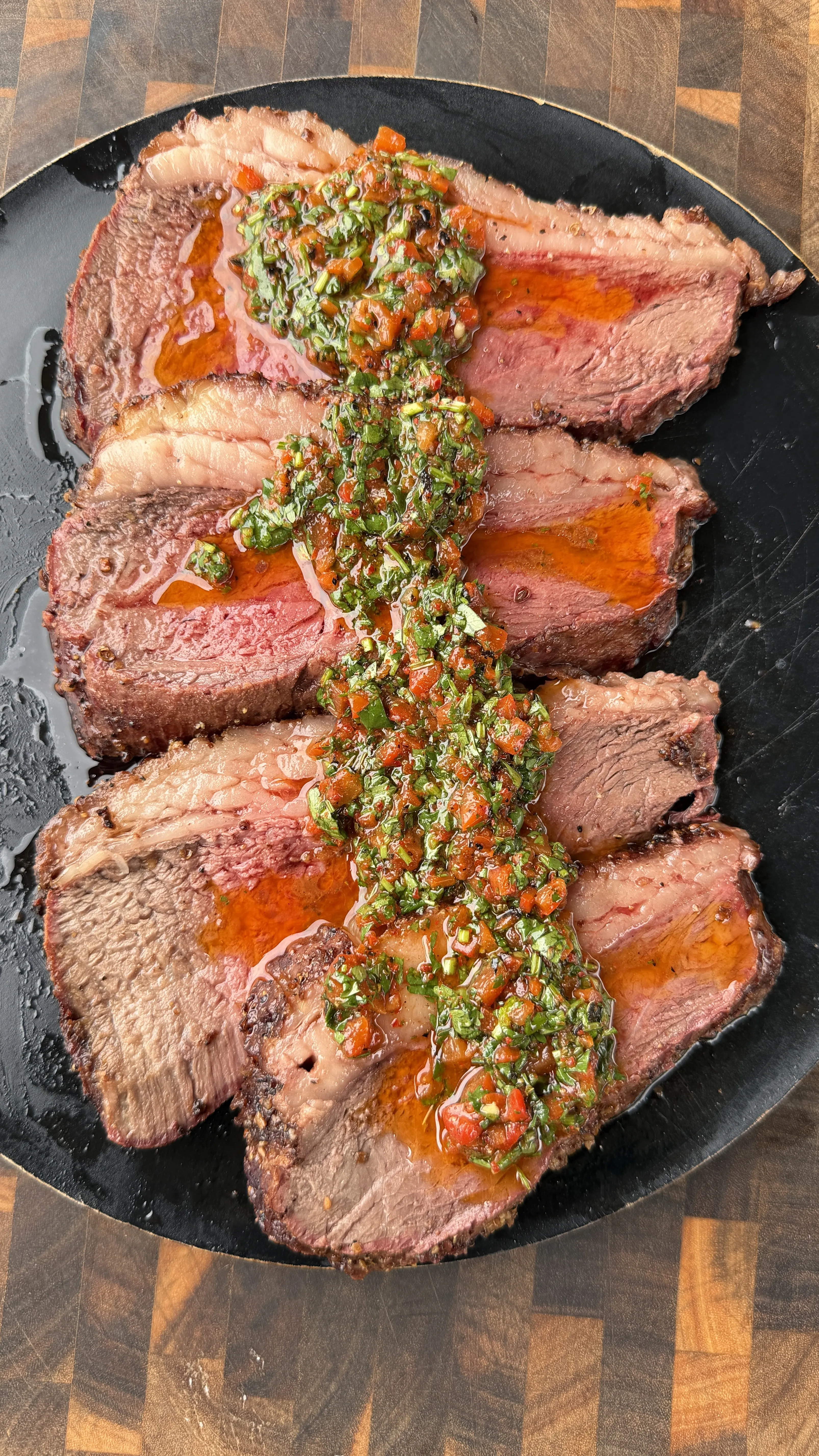 Sliced smoked picanha roast topped with red cilantro chimichurri sauce on a black plate