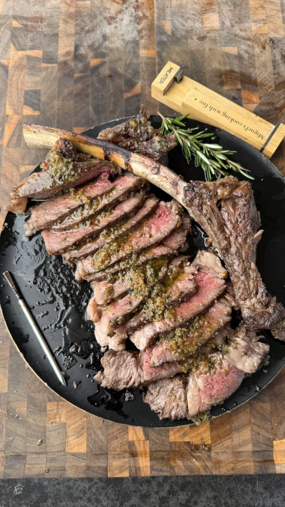 Sliced tomahawk ribeye steak topped with chimichurri sauce on a black cast iron plate with fresh rosemary