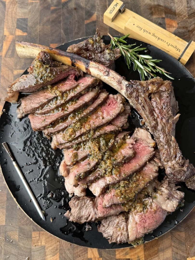 Sliced tomahawk ribeye steak topped with chimichurri sauce on a black cast iron plate with fresh rosemary