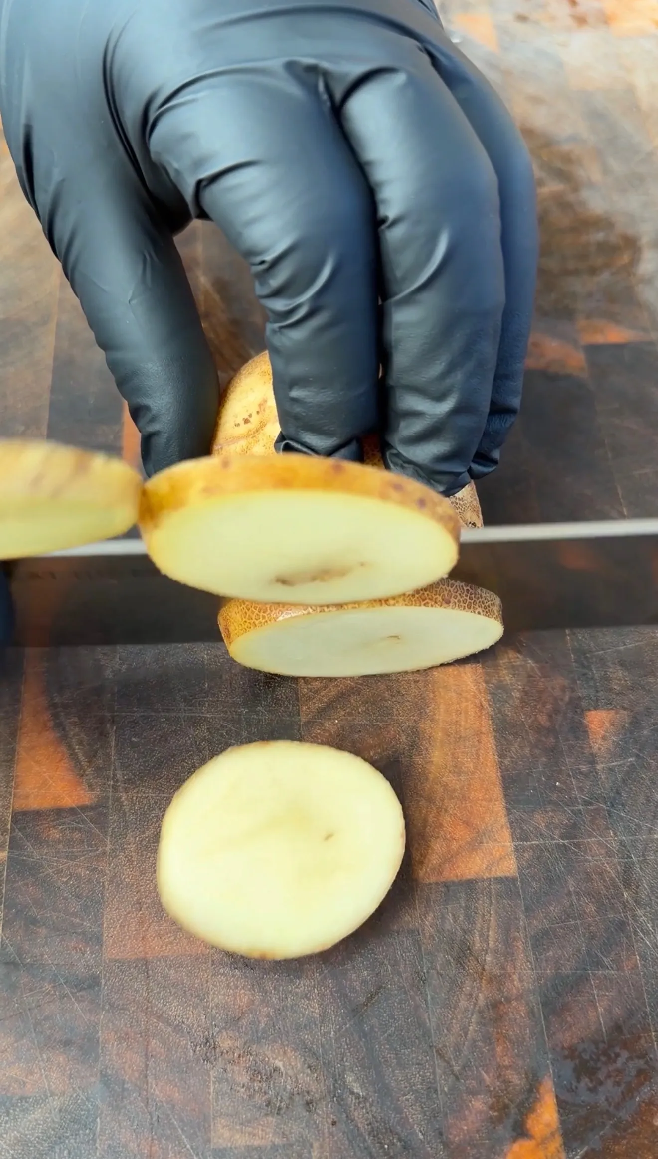 Gloved hands slicing raw potatoes into rounds on a dark wooden cutting board for Cajun beef kebabs