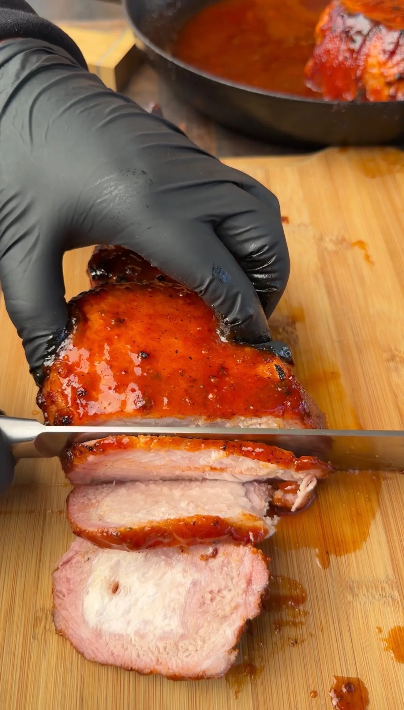 Gloved hands slicing a glazed smoked tomahawk pork chop with black raspberry BBQ sauce on a wooden cutting board
