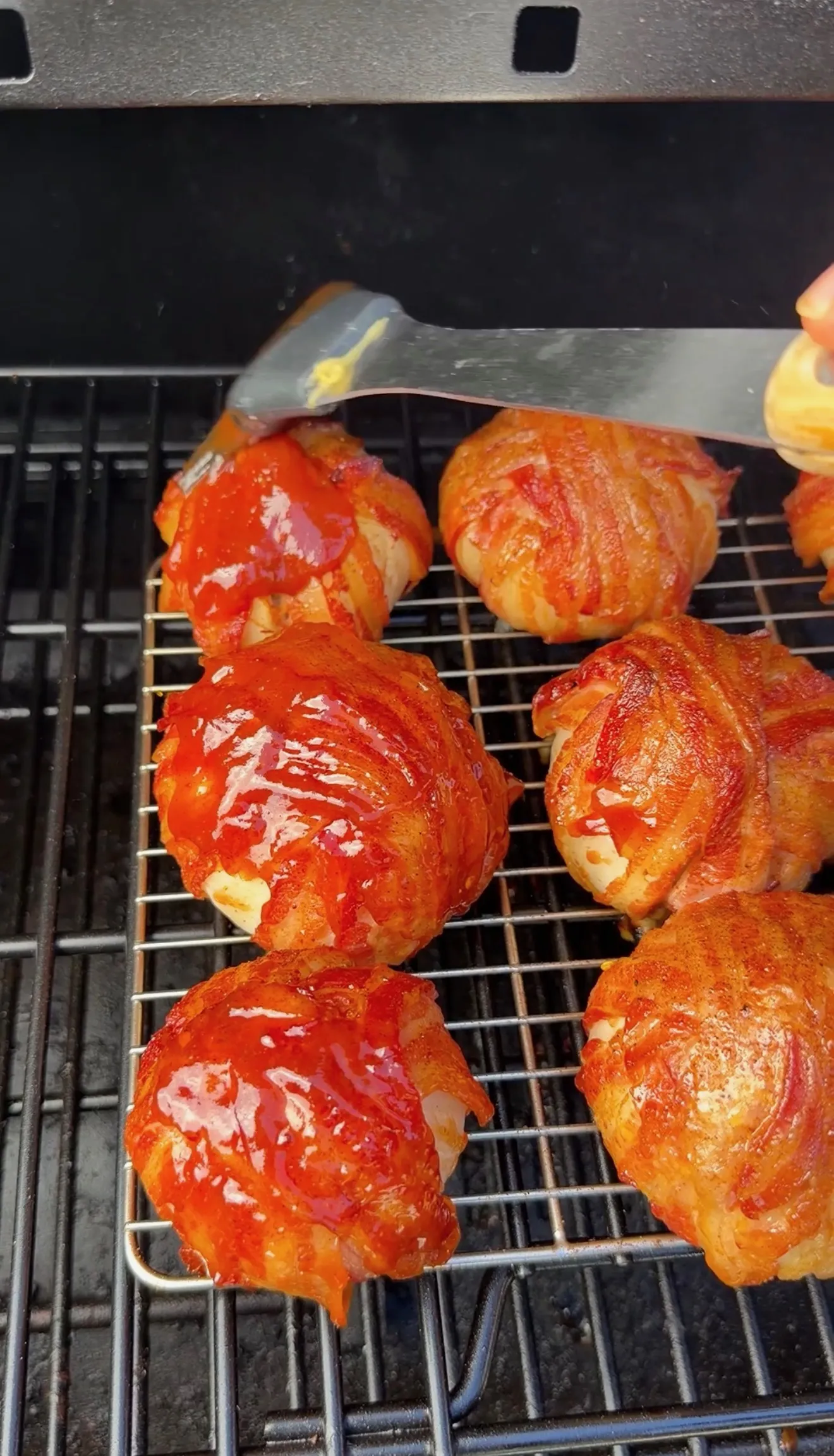 Bacon-wrapped onion bombs glazed with BBQ sauce cooking on smoker grill grates, being basted with spatula