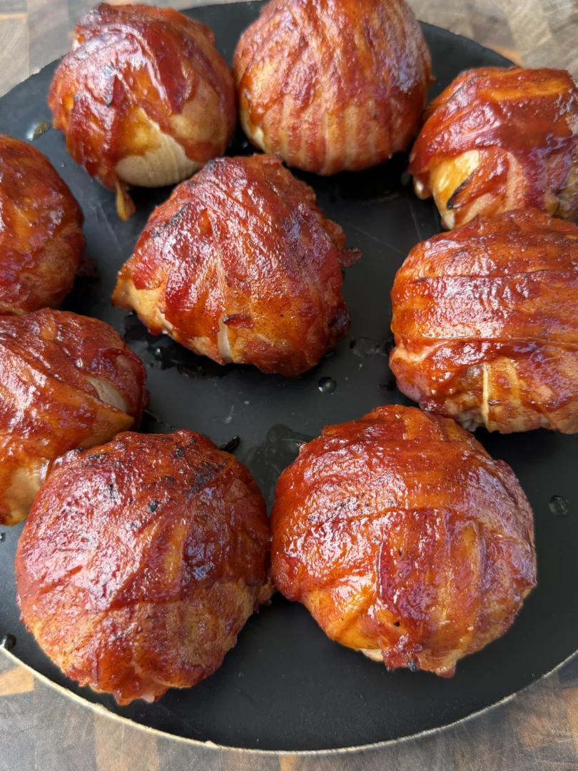 Nine BBQ-glazed smoked bacon-wrapped onion bombs arranged on a dark round plate on a wooden cutting board