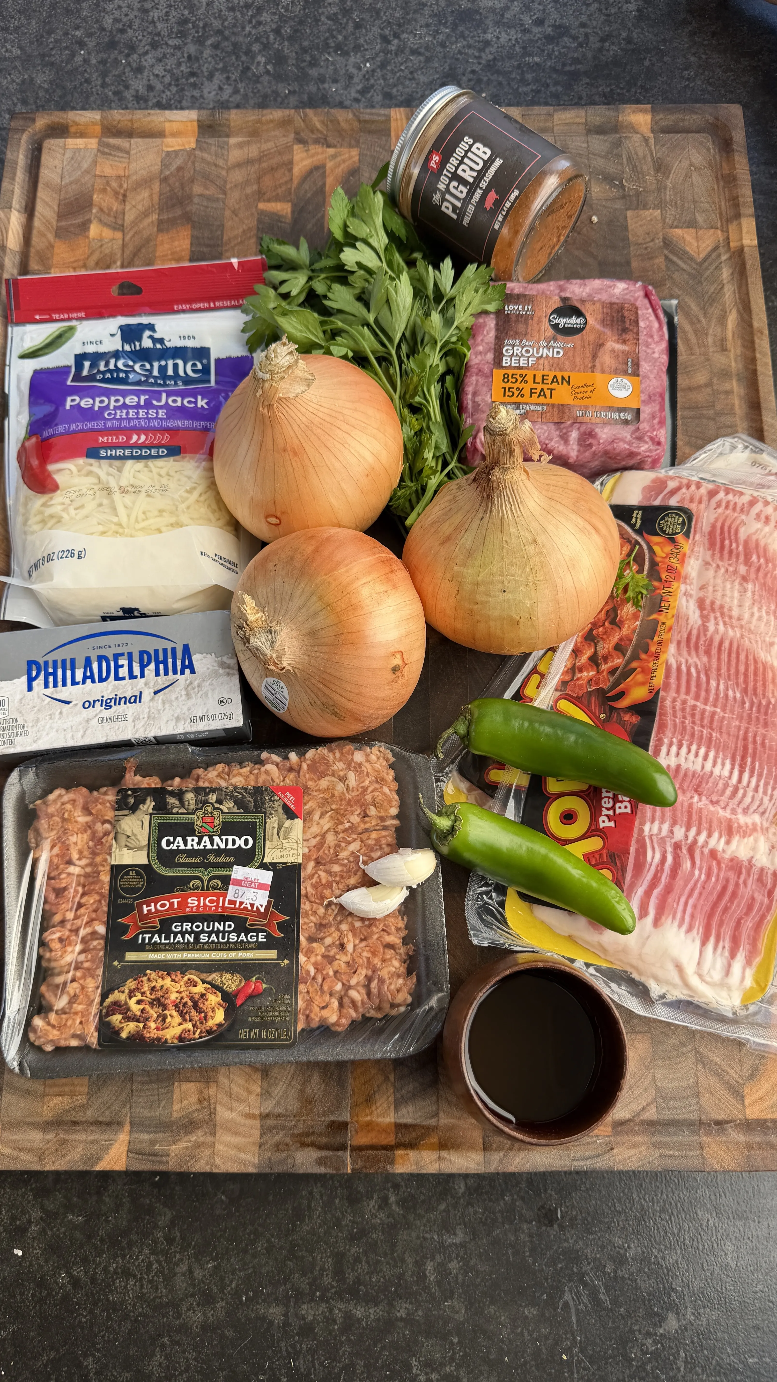 Ingredients for smoked bacon-wrapped onion bombs including ground beef, sausage, onions, cream cheese, jalapeños, and bacon on a wooden cutting board