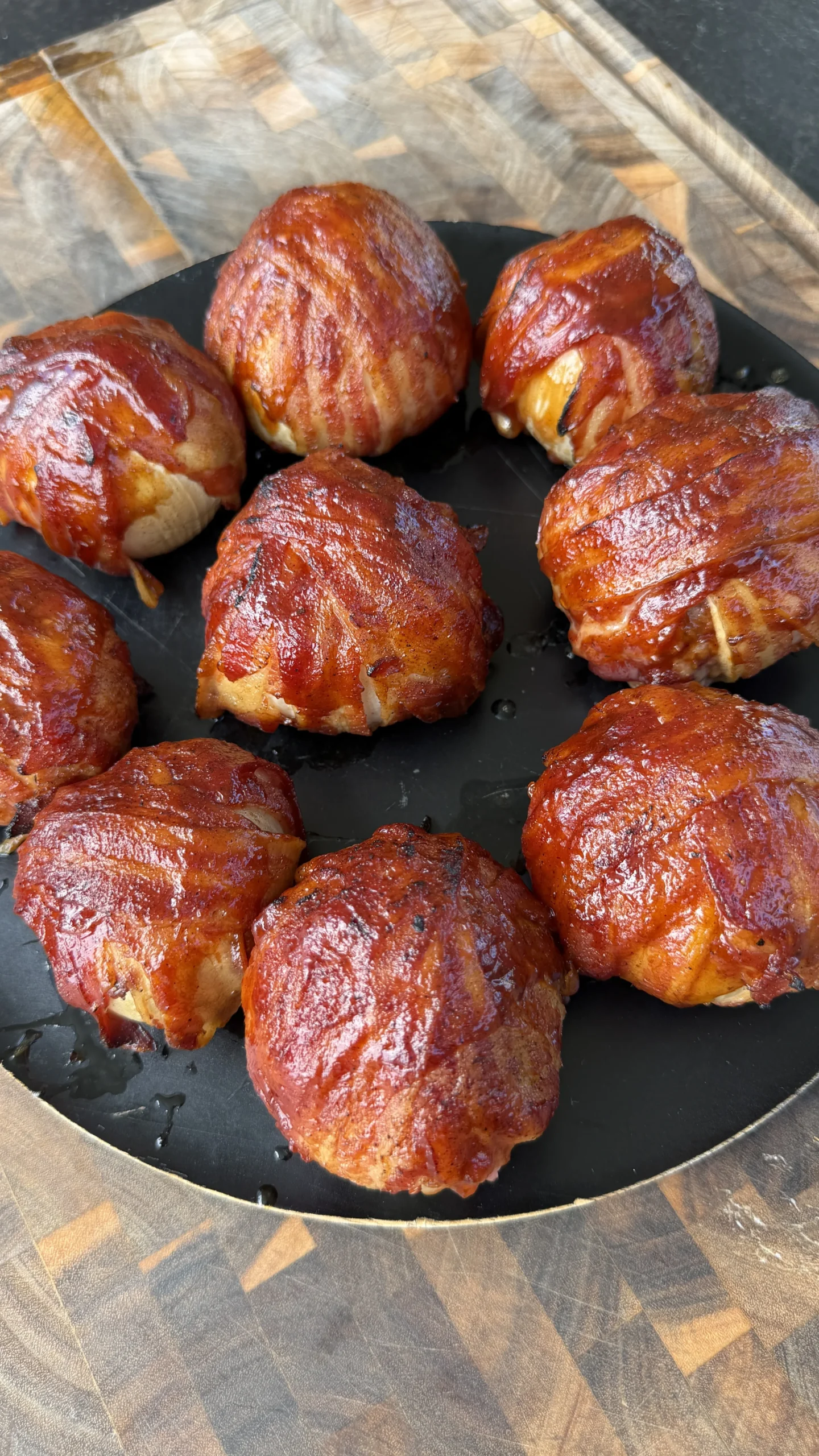 Eight glazed bacon-wrapped onion bombs arranged on a black round plate over a wooden cutting board