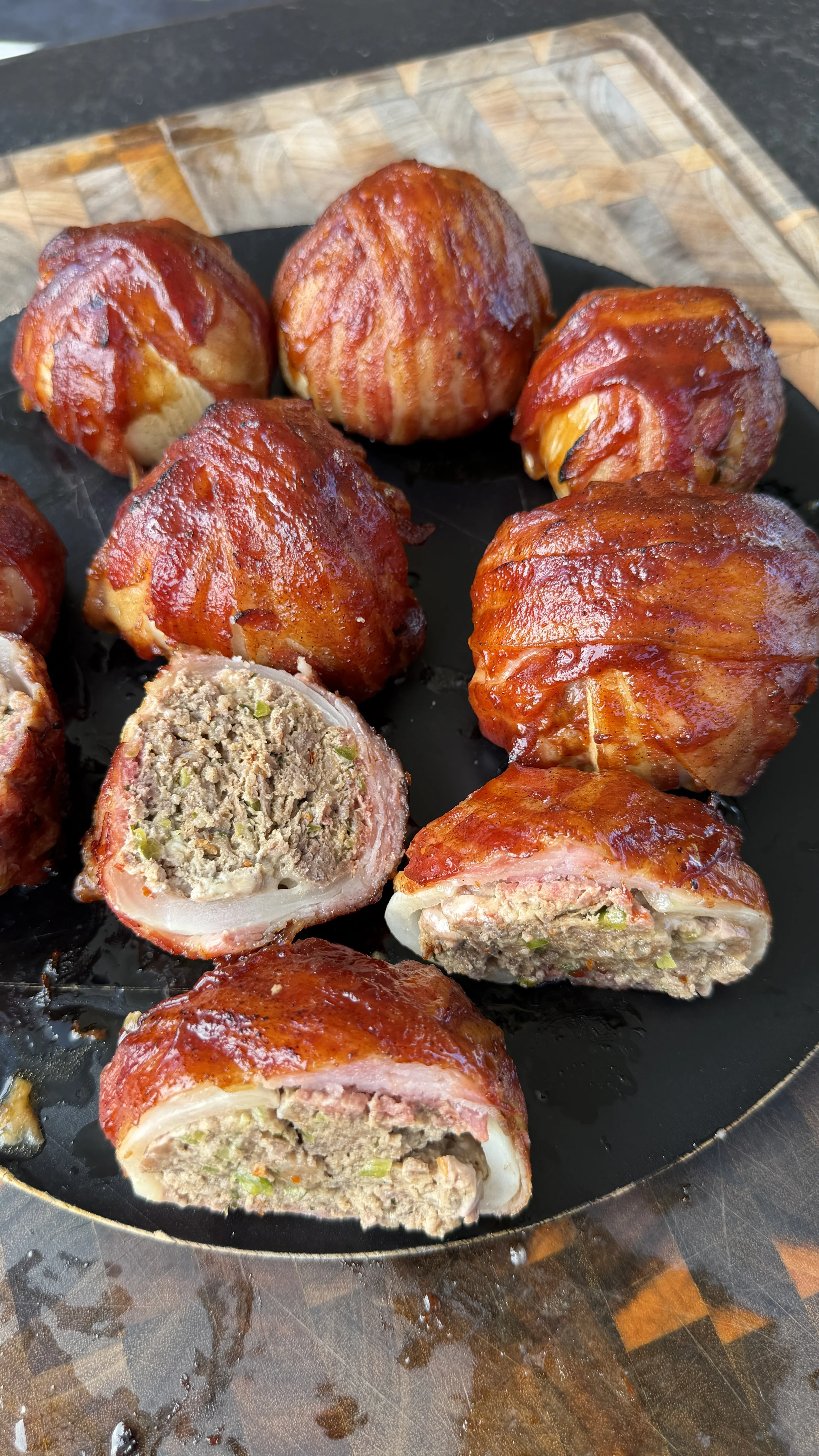 Bacon-wrapped onion bombs glazed with BBQ sauce on a black plate, two cut open revealing the seasoned meat filling