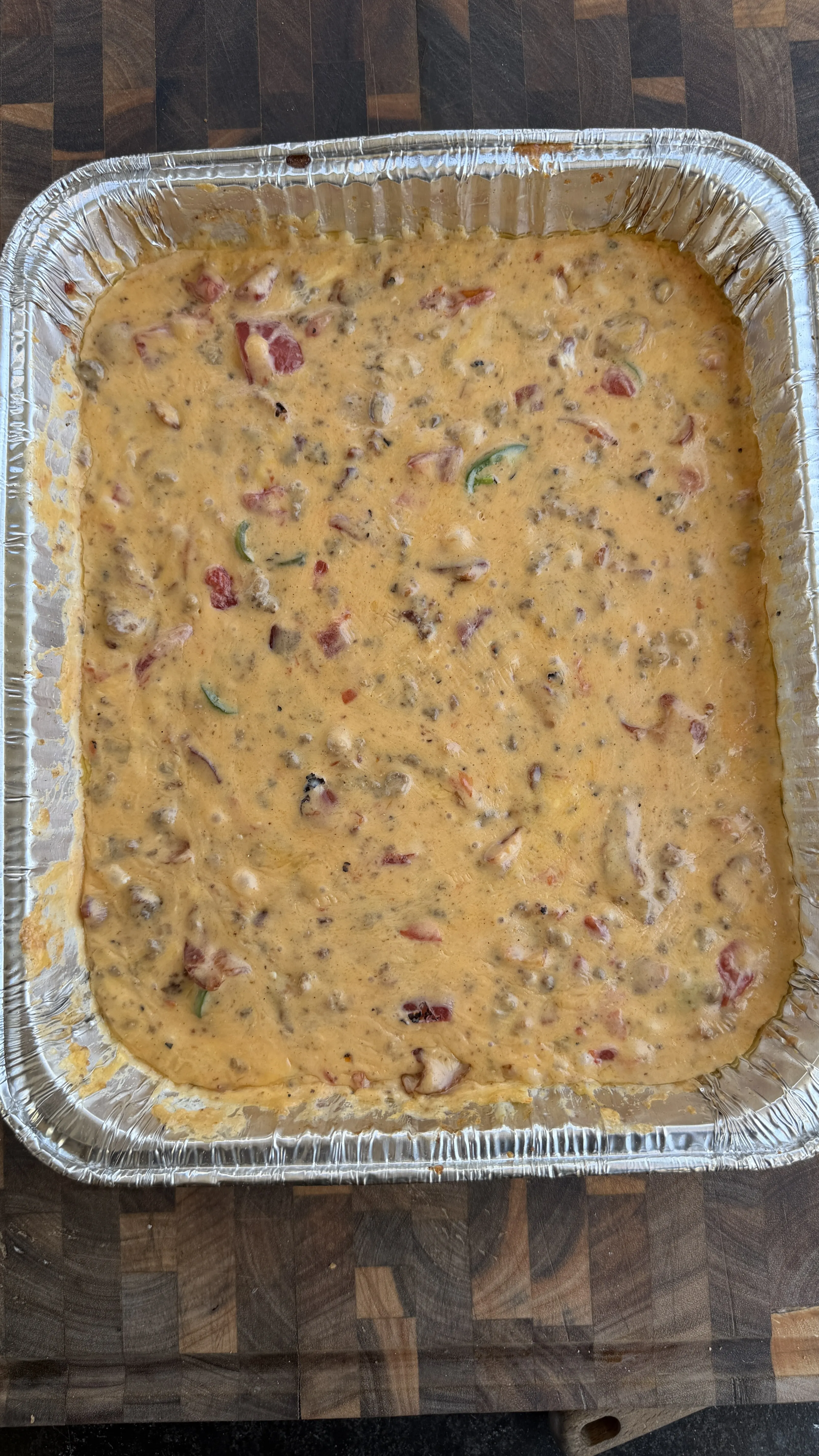 Creamy smoked cheeseburger queso dip with jalapeños and tomatoes in a large aluminum foil pan