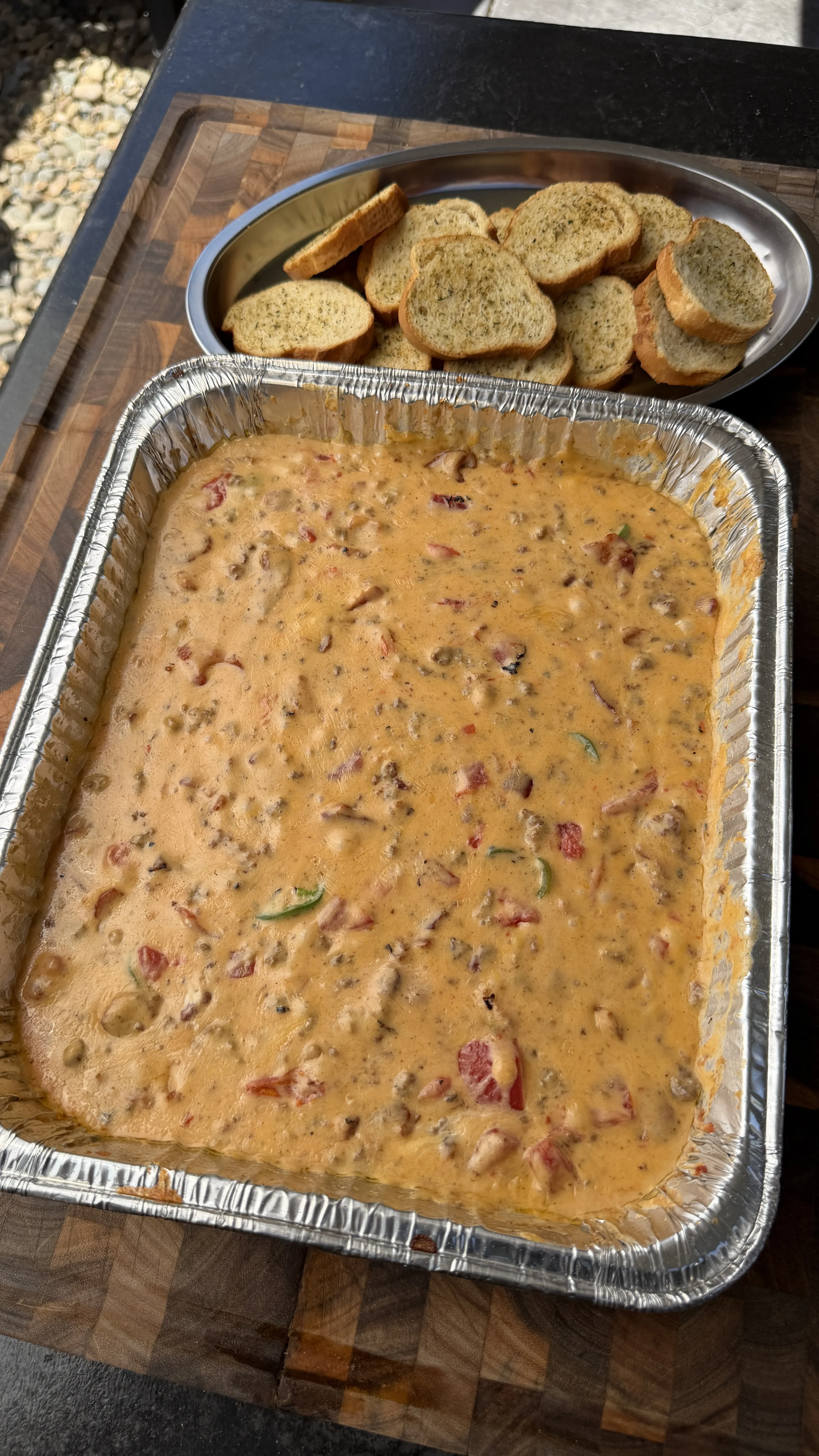 Creamy smoked cheeseburger queso dip in a foil pan with toasted crostini on a wooden cutting board