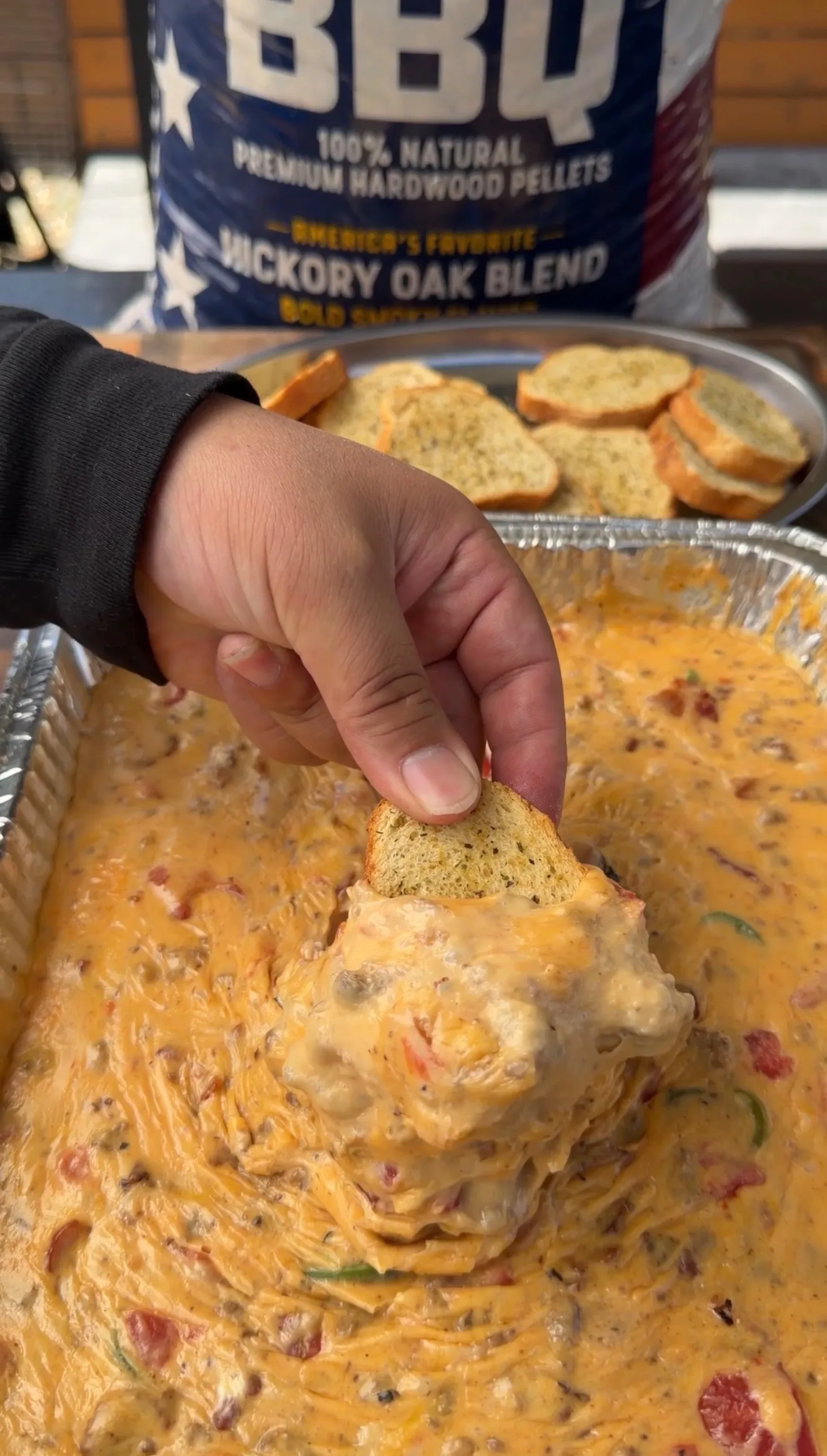 Hand dipping toasted bread into creamy smoked cheeseburger queso dip in aluminum pan with crostini in background