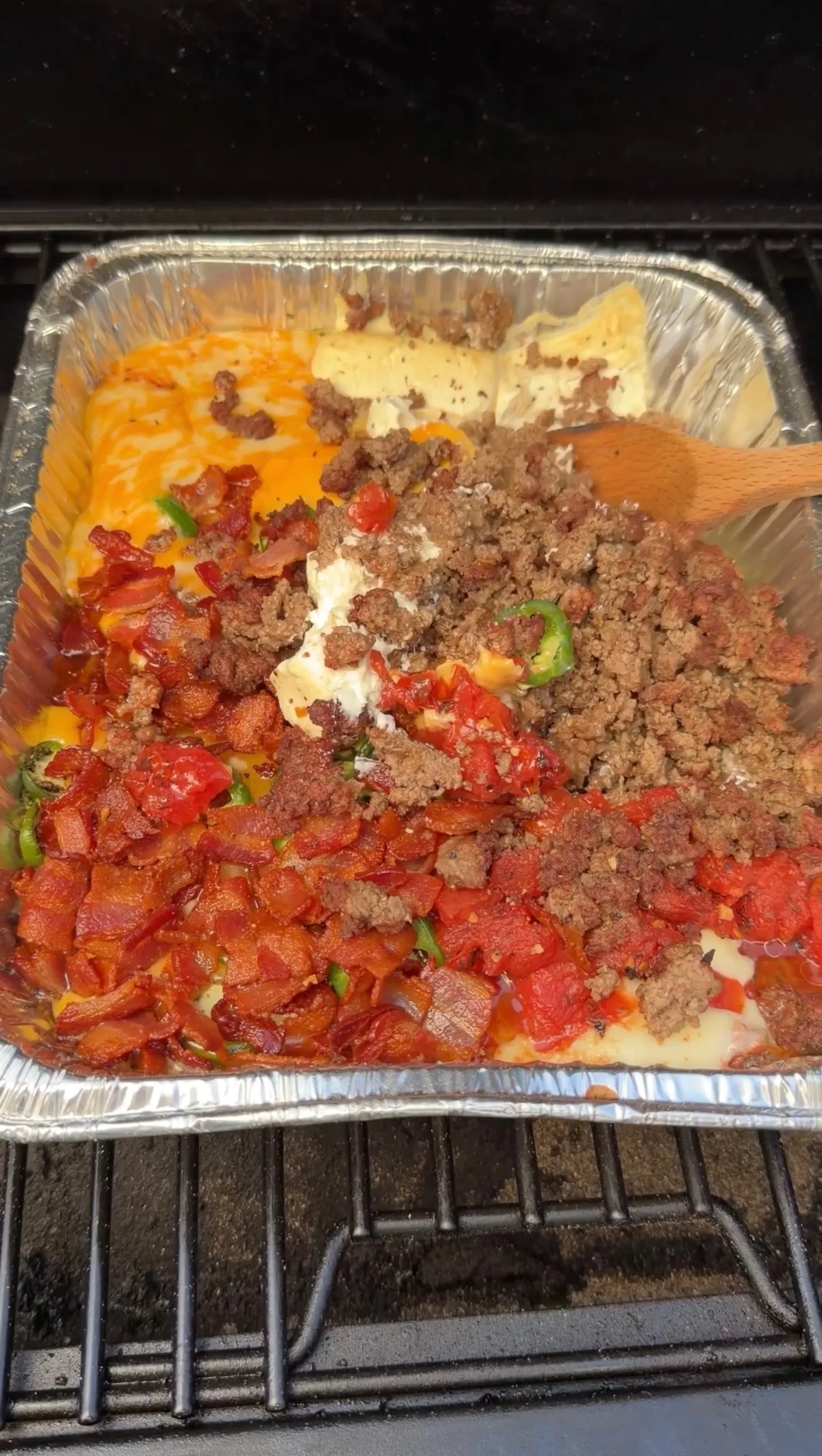 Smoked cheeseburger queso dip being mixed in a foil pan with ground beef, bacon, tomatoes, and melted cheese