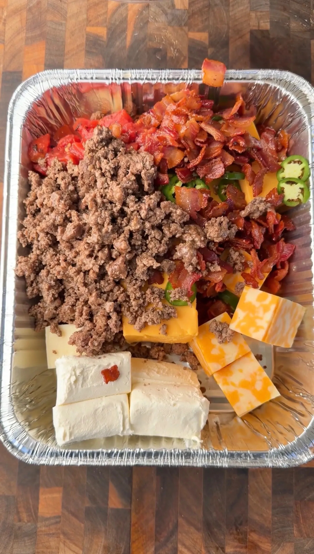 Raw ingredients for smoked cheeseburger queso dip in foil pan: ground beef, bacon, cream cheese, cheddar, jalapeños, tomatoes