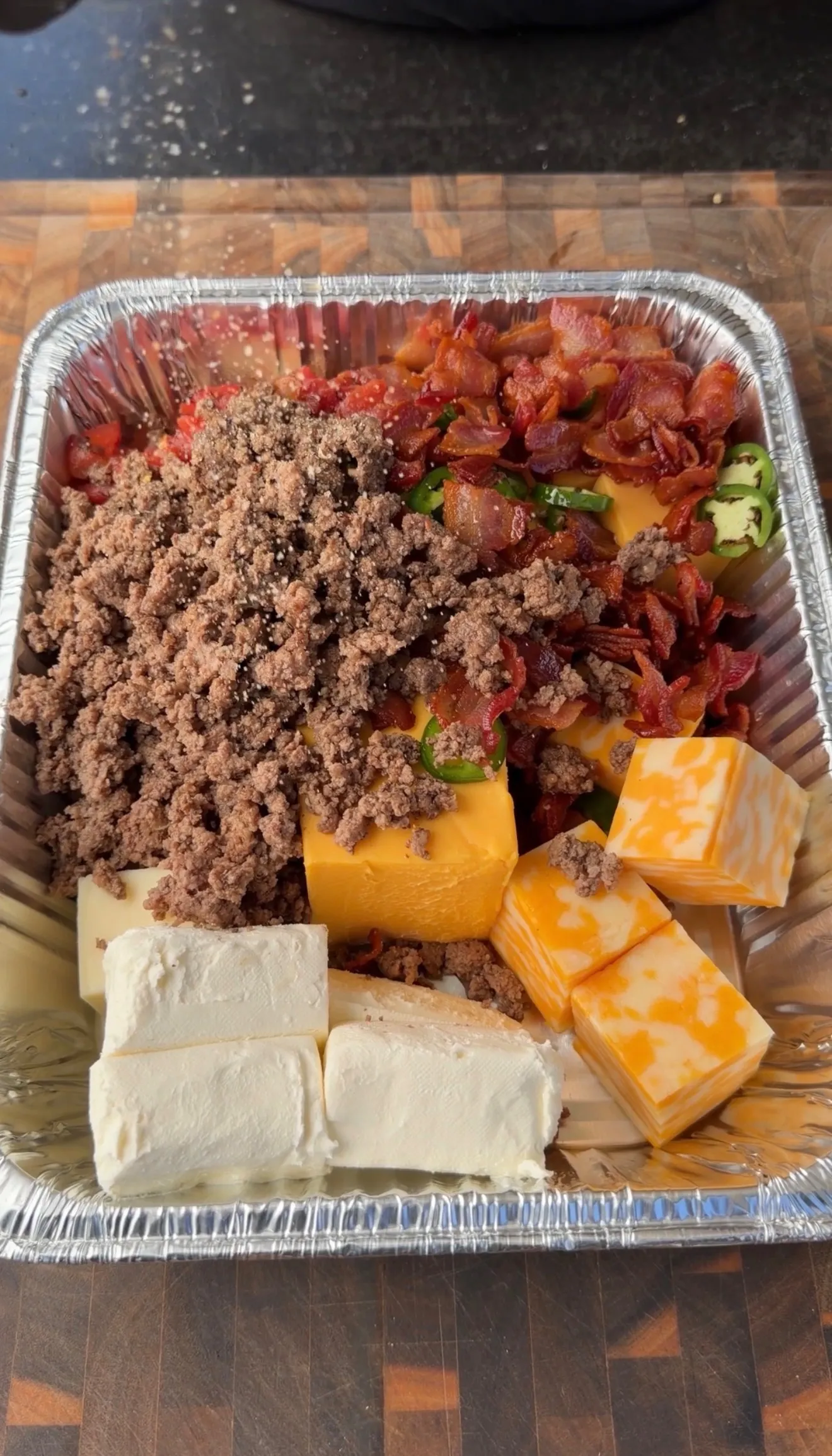 Aluminum pan with ground beef, cream cheese, cheddar, Velveeta, bacon, jalapeños, and tomatoes for smoked queso dip
