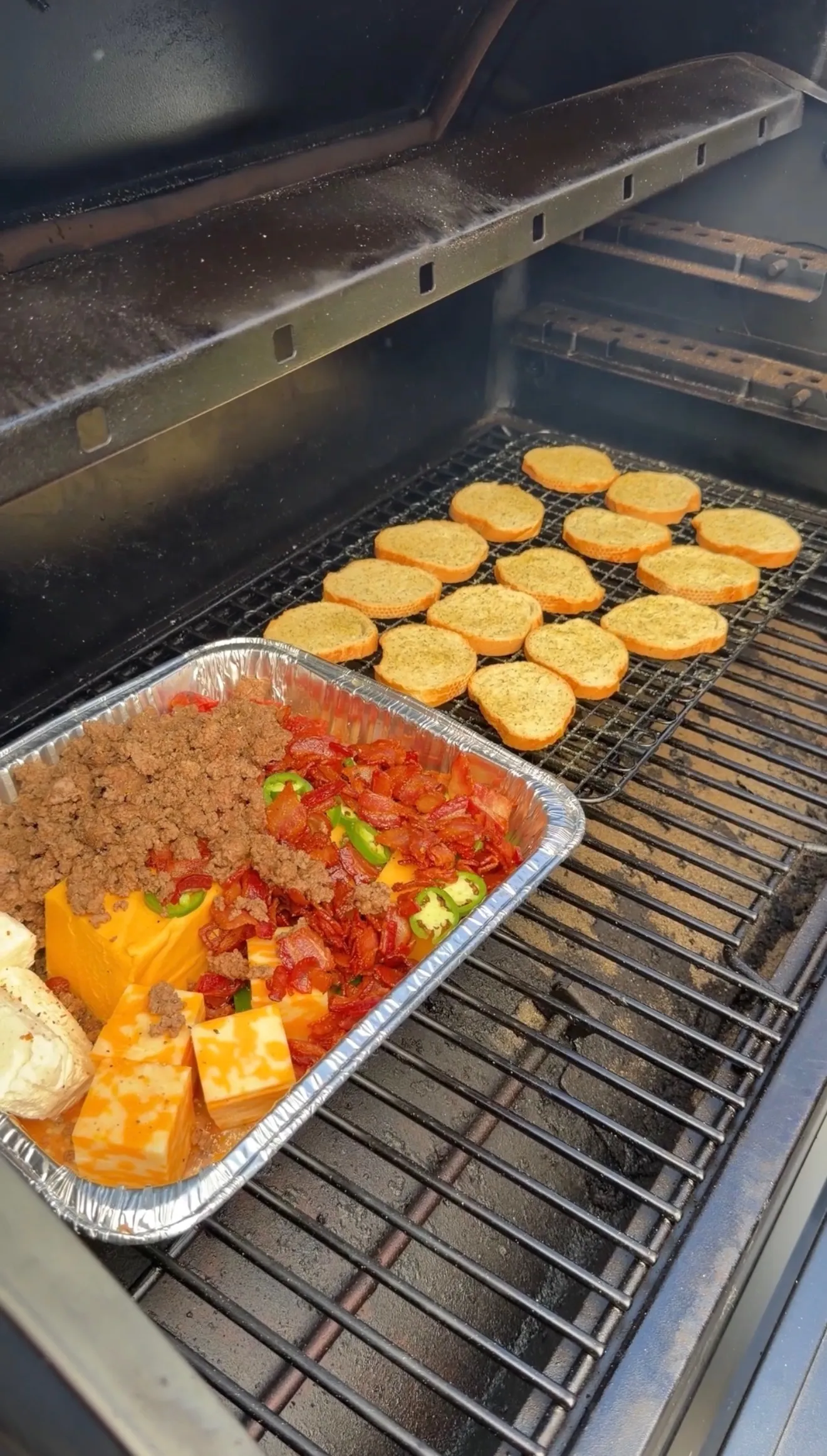 Smoked cheeseburger queso dip with Velveeta, bacon, jalapeños, and ground beef in foil pan beside toasted buns on smoker grill