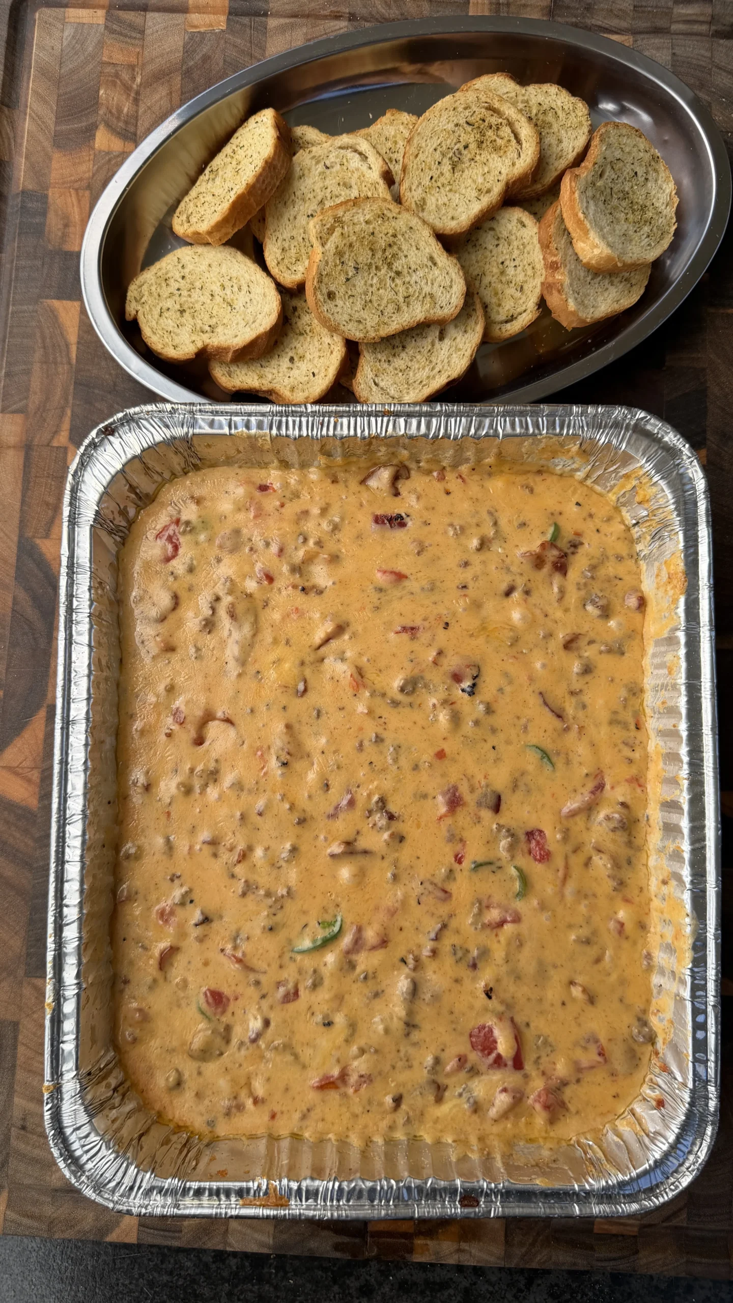 Smoked cheeseburger queso dip in a foil pan with melted cheese, meat, and peppers, served with toasted crostini