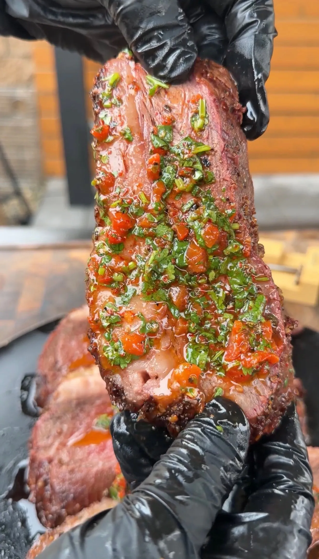 Hands holding a smoked picanha roast topped with vibrant red and green cilantro chimichurri sauce