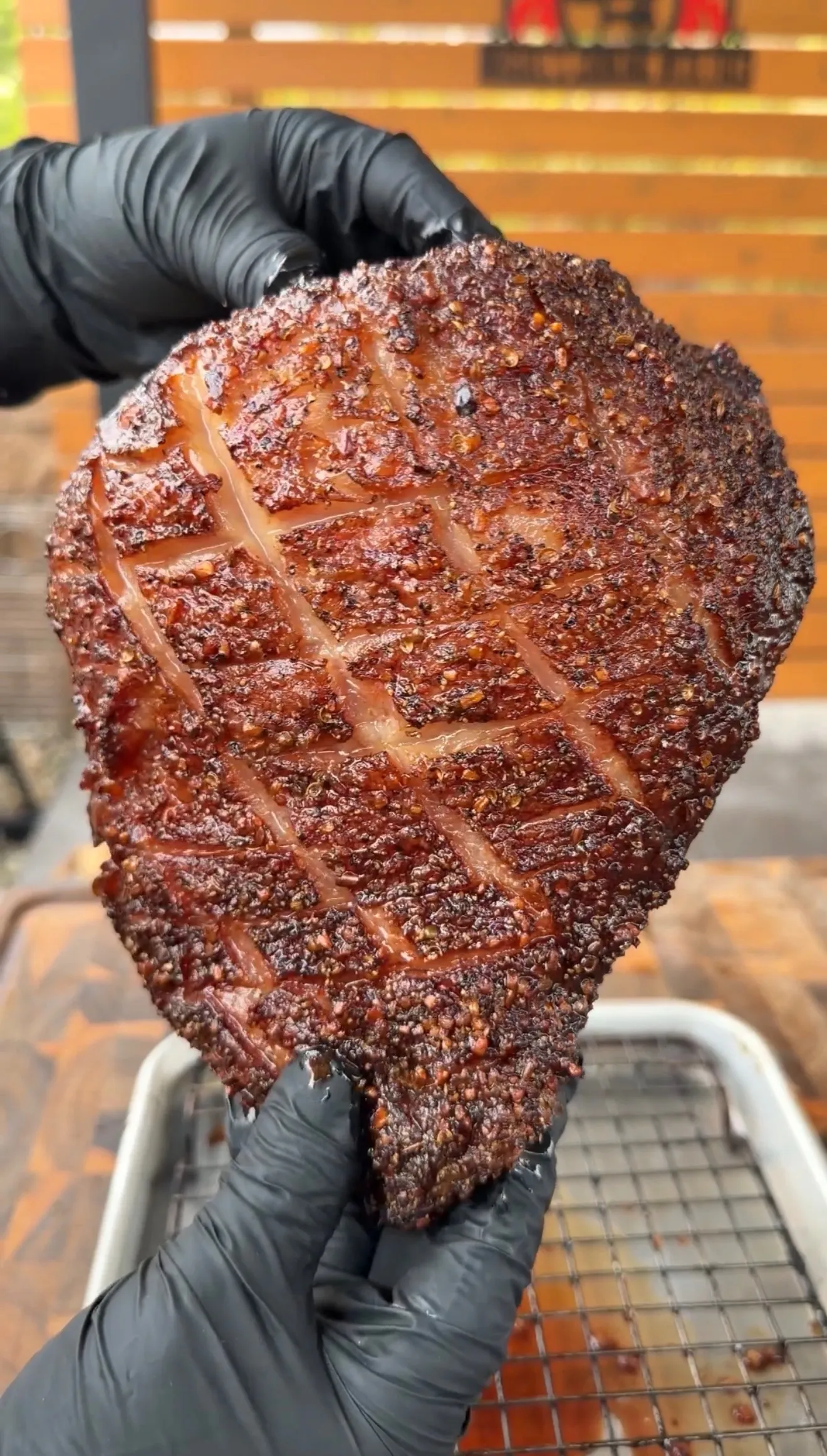 Hands in black gloves holding a smoked picanha roast with a deeply caramelized, cross-hatched scored fat cap